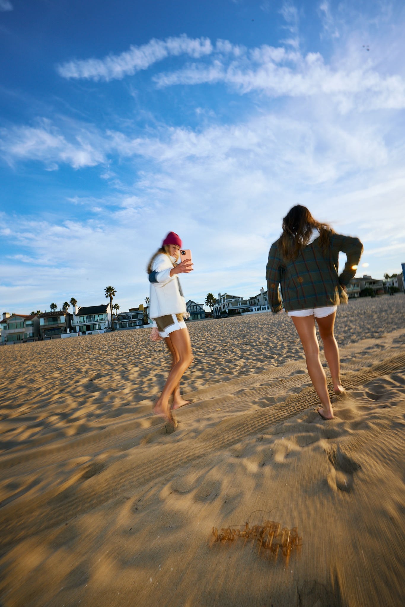 Two young women walking on a sandy beach during sunset or late afternoon, with beach houses in the background, one taking a selfie.