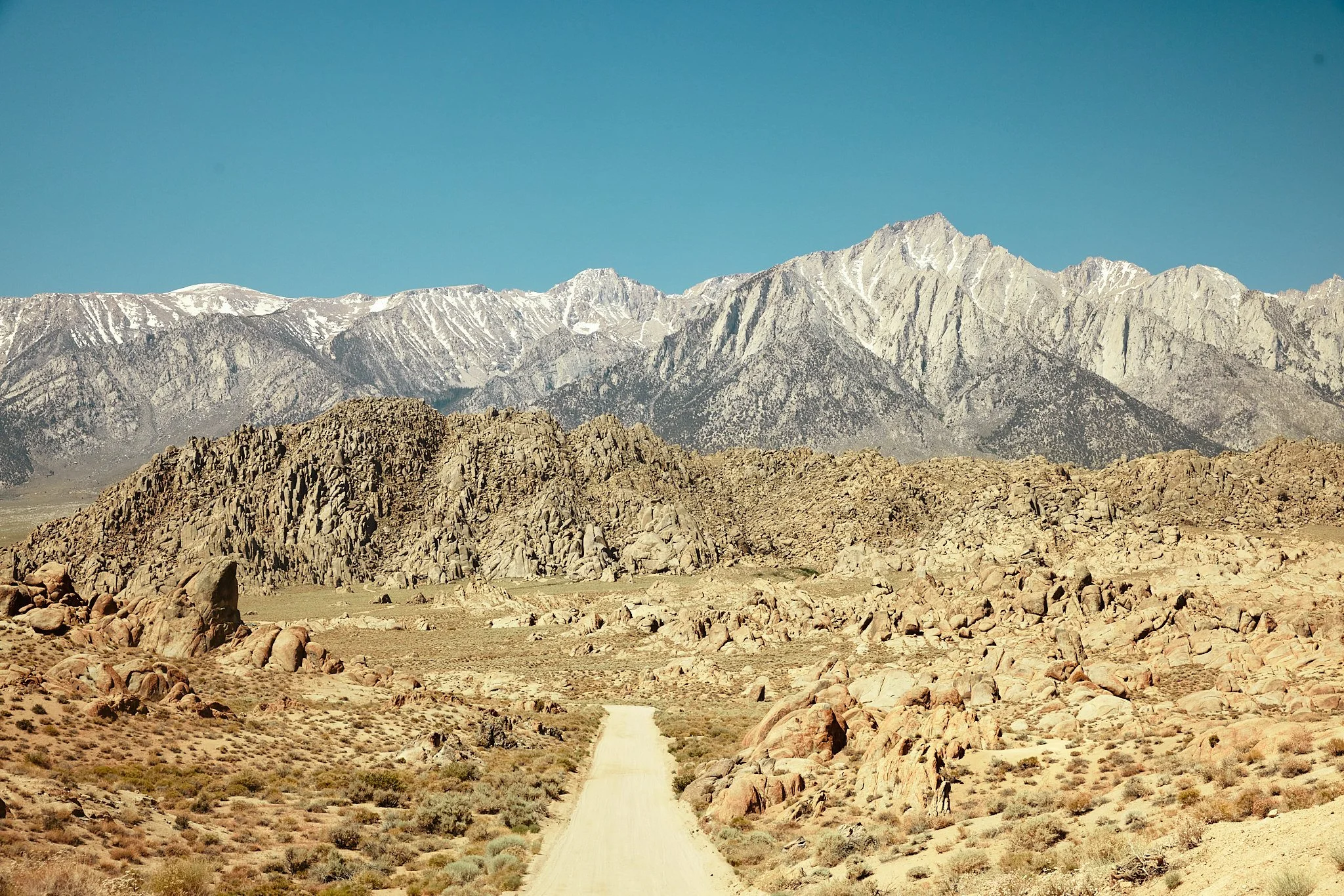 A dirt road running through a desert landscape with rocky terrain, leading towards snow-capped mountains under a clear blue sky.