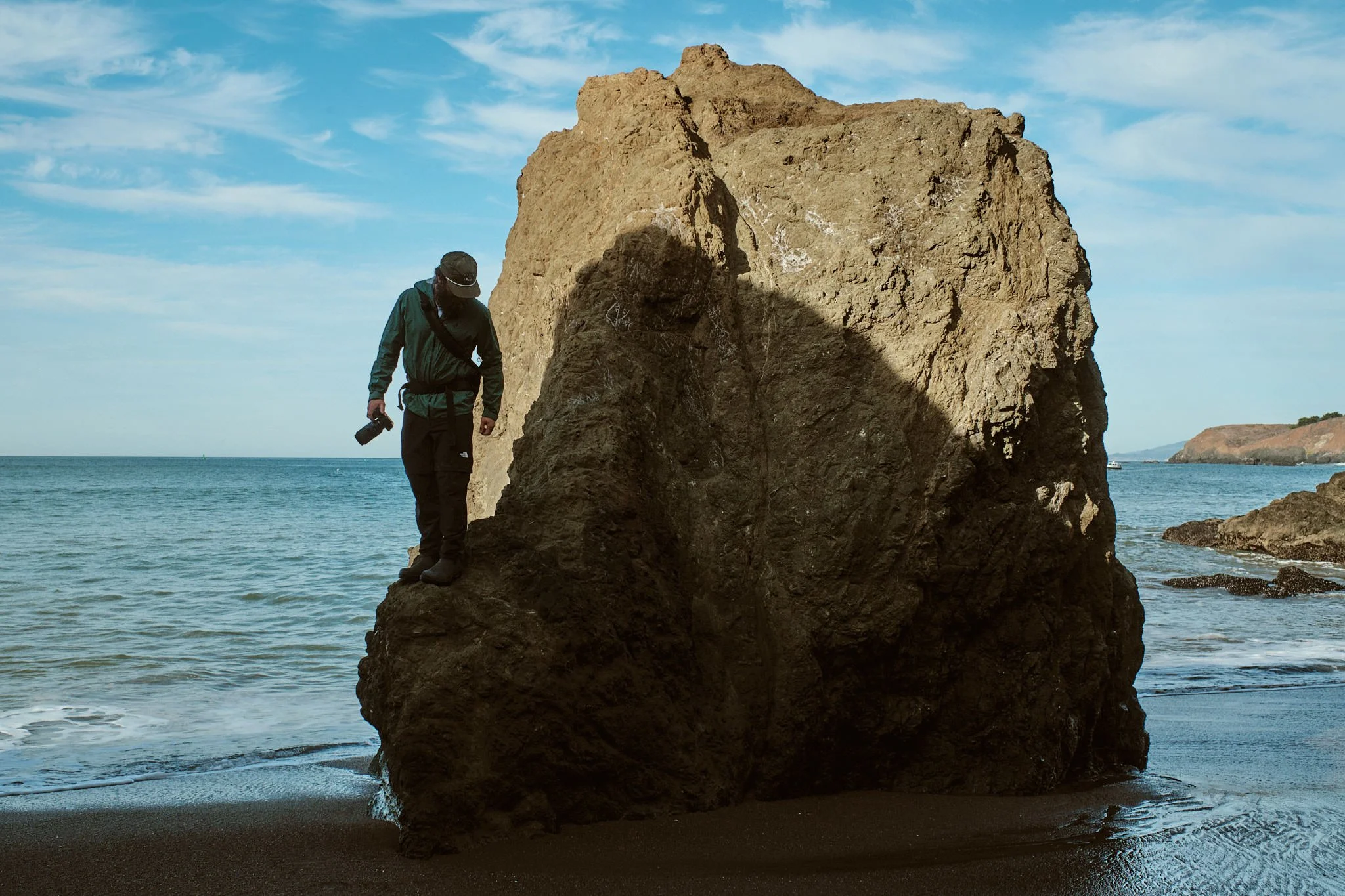 A person standing on a beach, looking at a large rock formation near the shoreline under a partly cloudy sky.