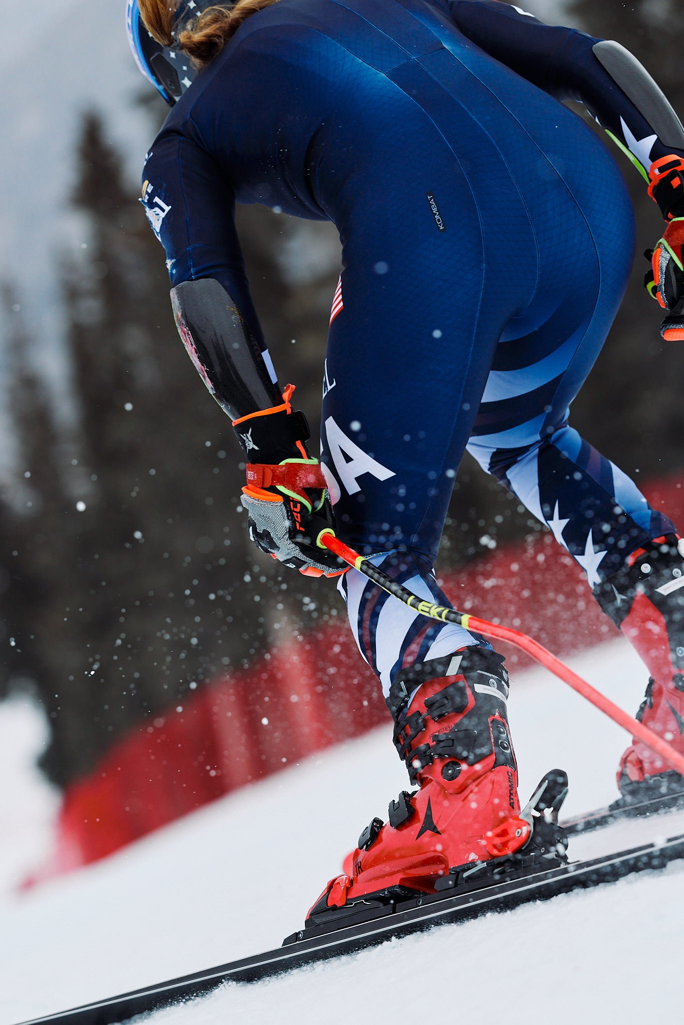 A skier wearing a blue suit and red ski boots skiing down a snowy slope with snow-covered trees in the background.
