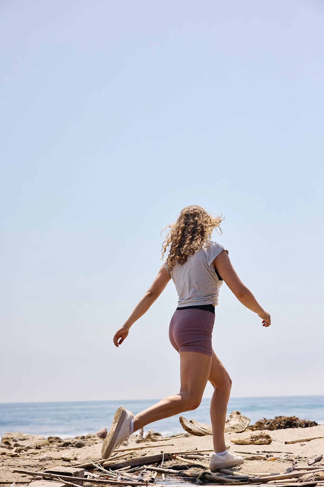 A woman with curly hair jogging on a rocky beach, wearing athletic clothes, with a clear blue sky and ocean in the background.