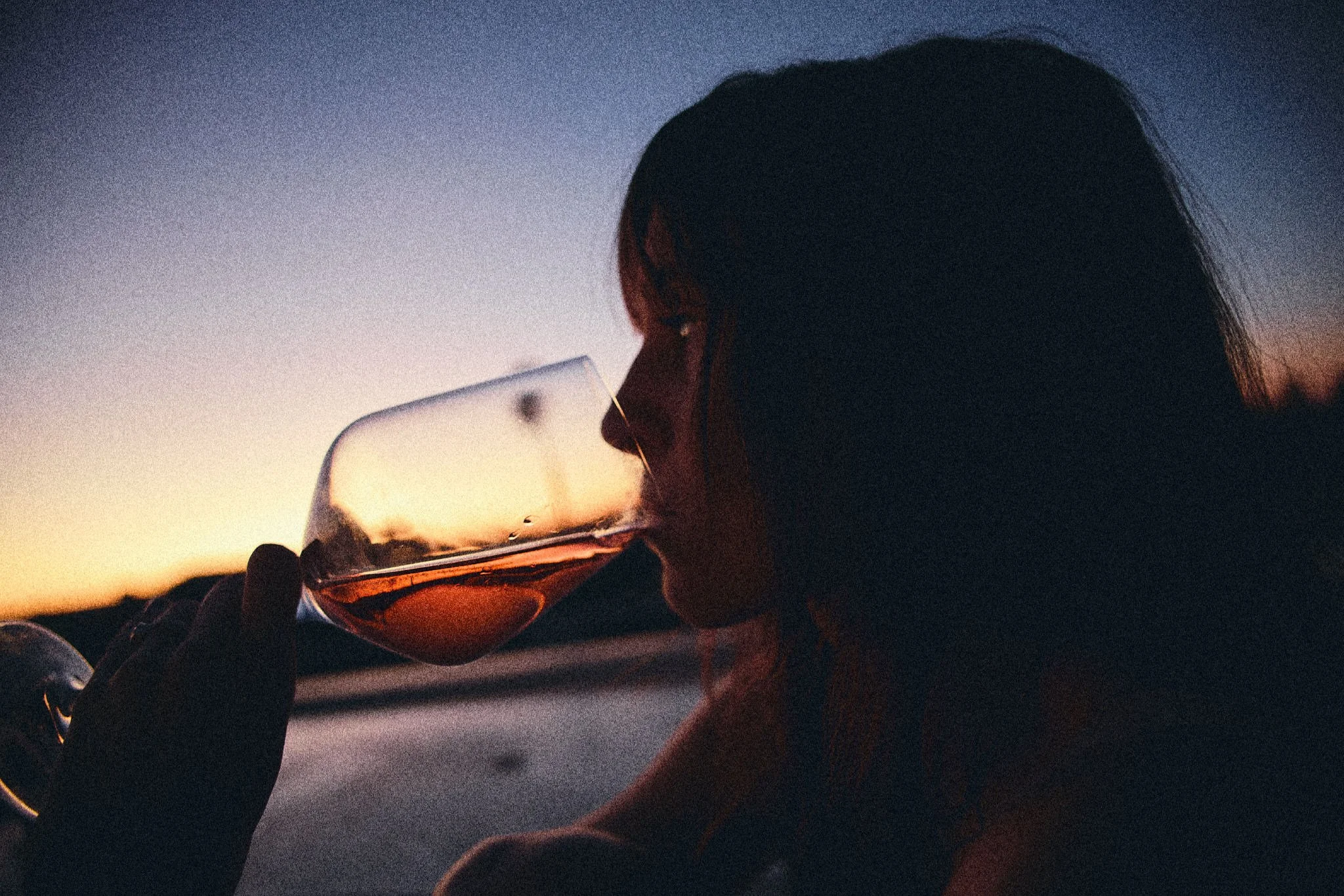 A person with long dark hair drinking wine from a glass at sunset.