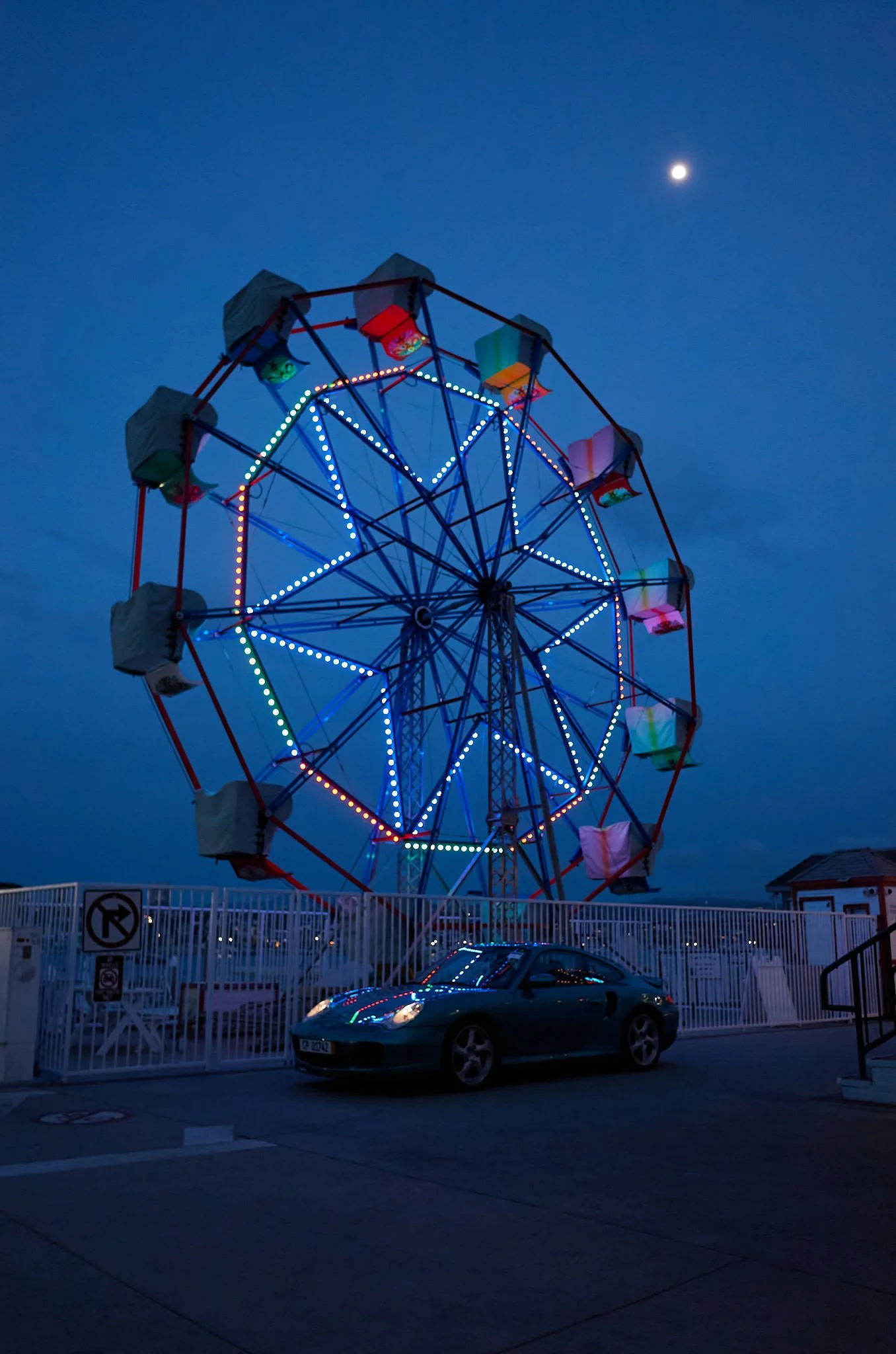 A large Ferris wheel with colorful lights is illuminated against a dark blue sky near the moon; a black car is parked in front of a white fence.