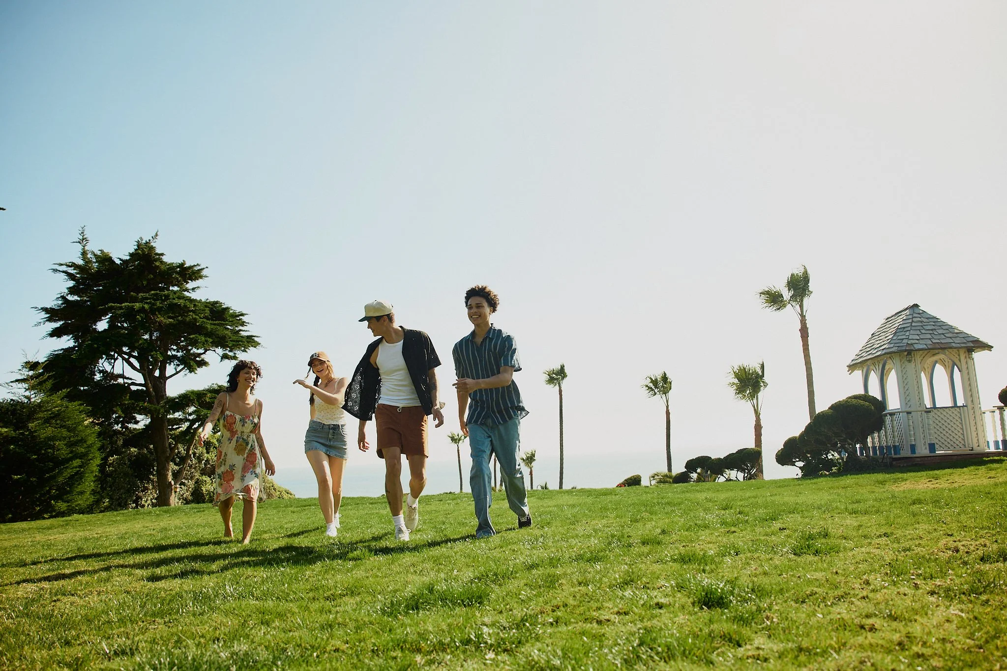 A group of five young people walking on a lush green lawn in a park on a sunny day. There are trees, palm trees, and a gazebo in the background.