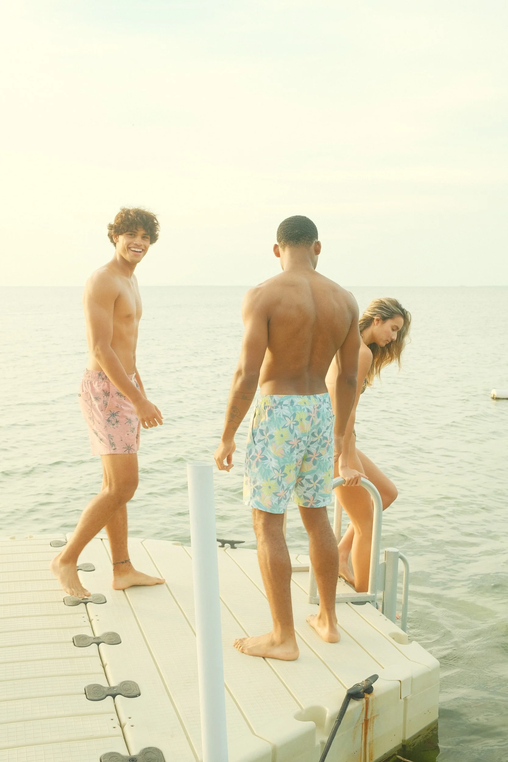 Three young adults in swimsuits on a dock by the water, with the ocean and sky in the background. One smiling person is to the left, another facing away, and a woman on the right is looking down.