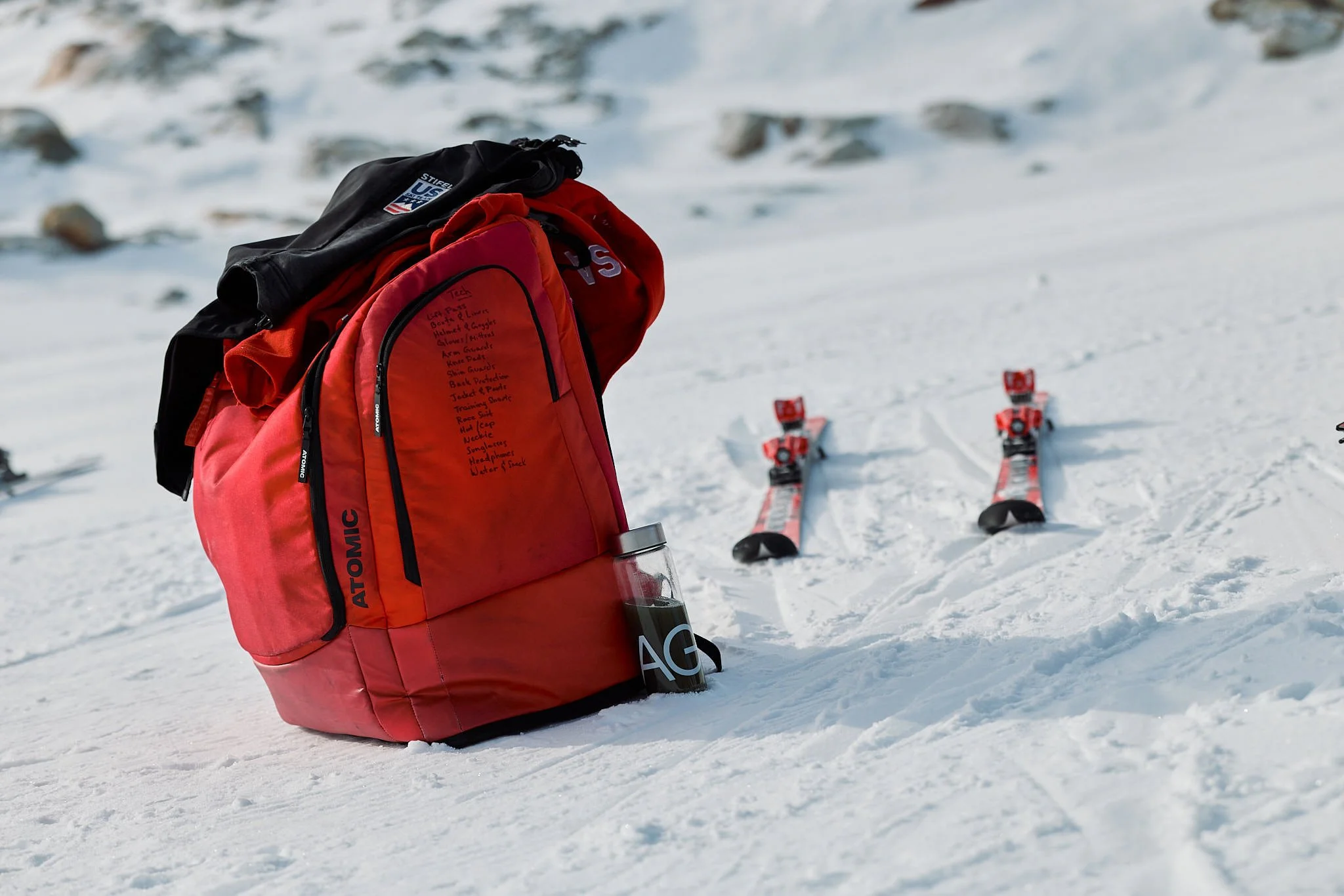 Red and black backpacks and a water bottle on snowy ground near a ski slope with red skis in the background.