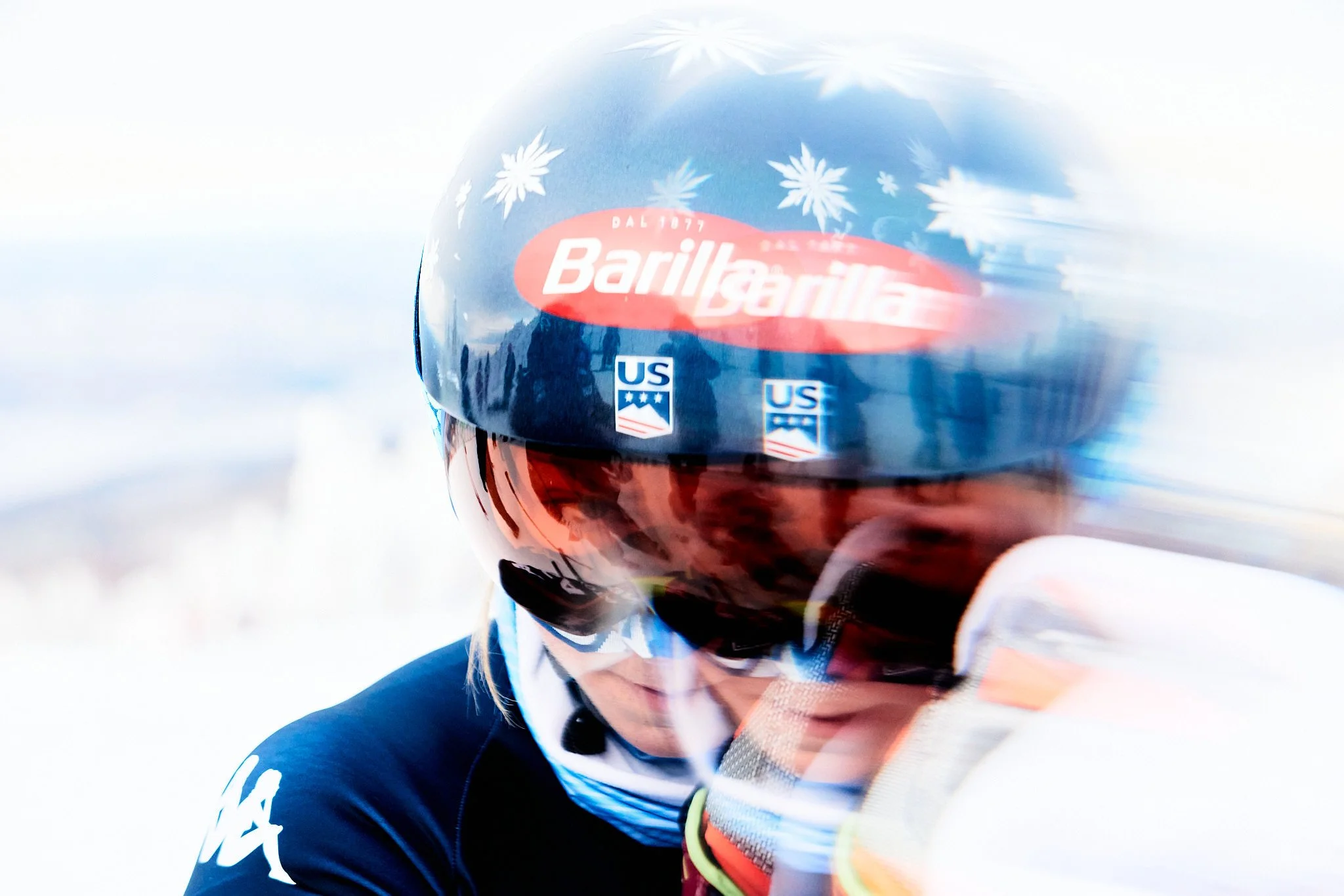 Close-up of a cyclist wearing a blue helmet with white snowflake patterns and a red logo, dressed in a dark racing suit with white and red accents, with a blurred background.