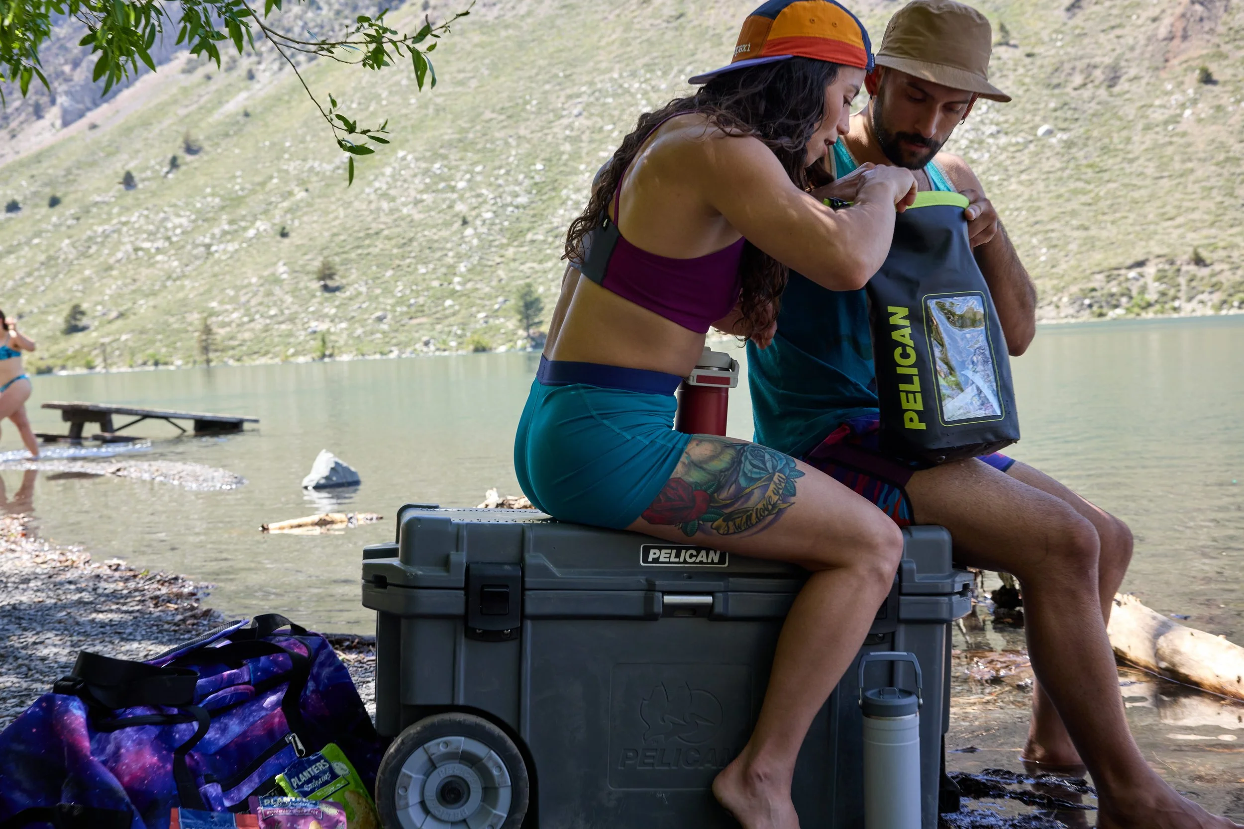 A man and woman sitting by a lake, packing or unpacking a backpack. The woman is wearing a purple top and blue shorts, has long curly hair, and tattoos on her thigh. The man is wearing a hat and a tank top. There are other backpacks and items around 