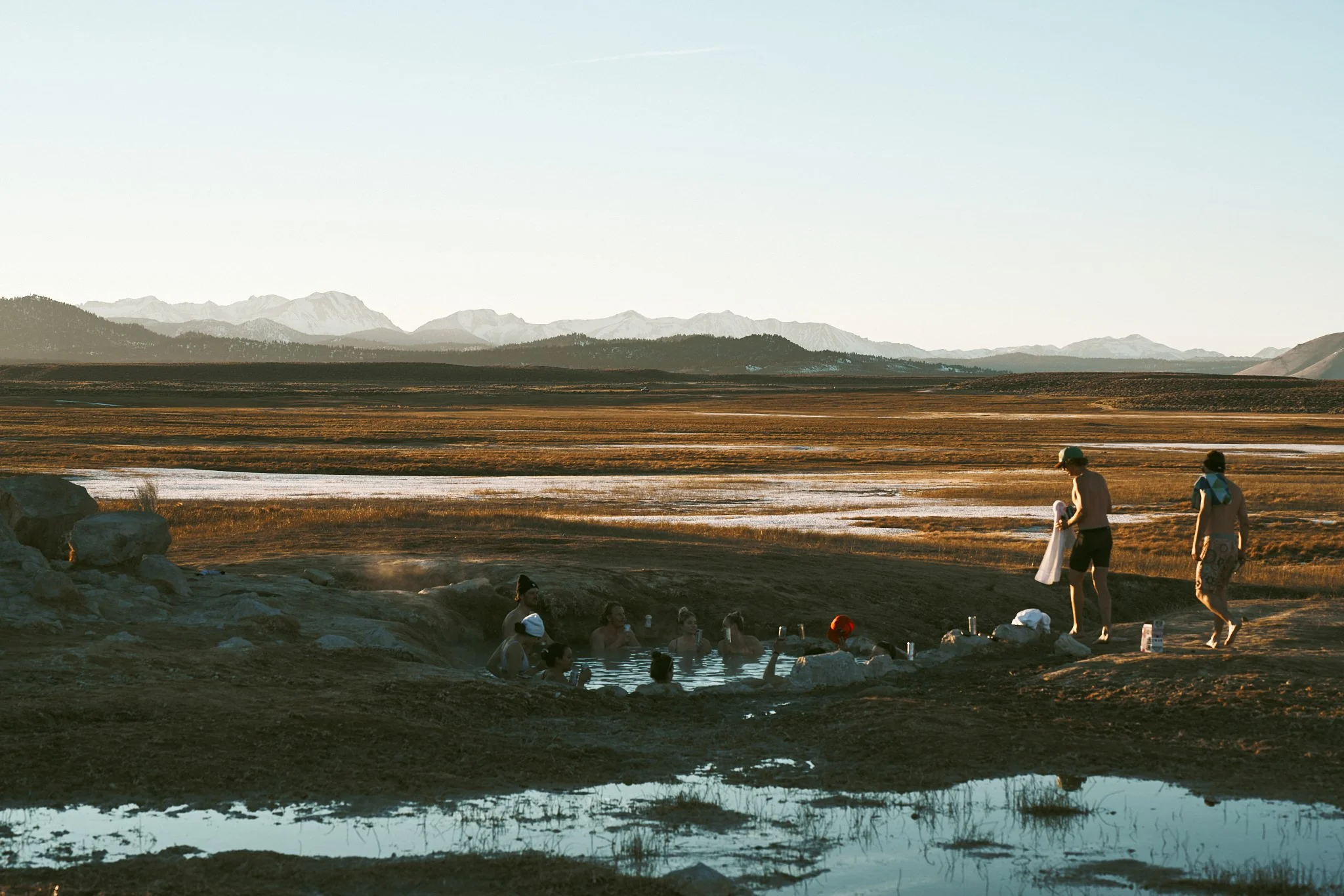 People enjoying a hot spring or geothermal pool outdoors with mountain range in the background during late afternoon or early evening.