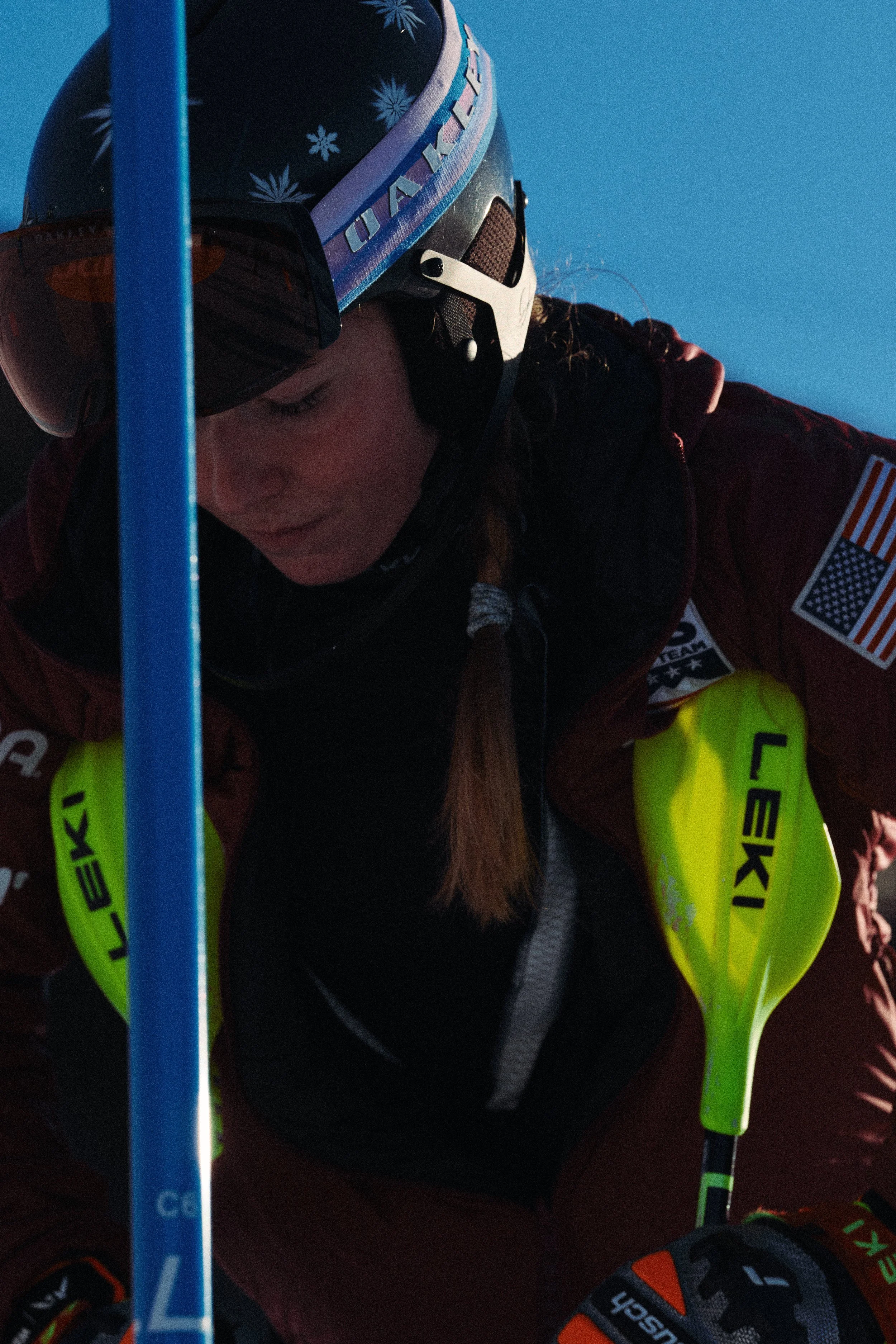 Close-up of a young woman in ski gear, wearing a black helmet with snowflake and snow pattern, a dark jacket with patches and ribbed collar, and bright yellow gloves, against a clear blue sky.