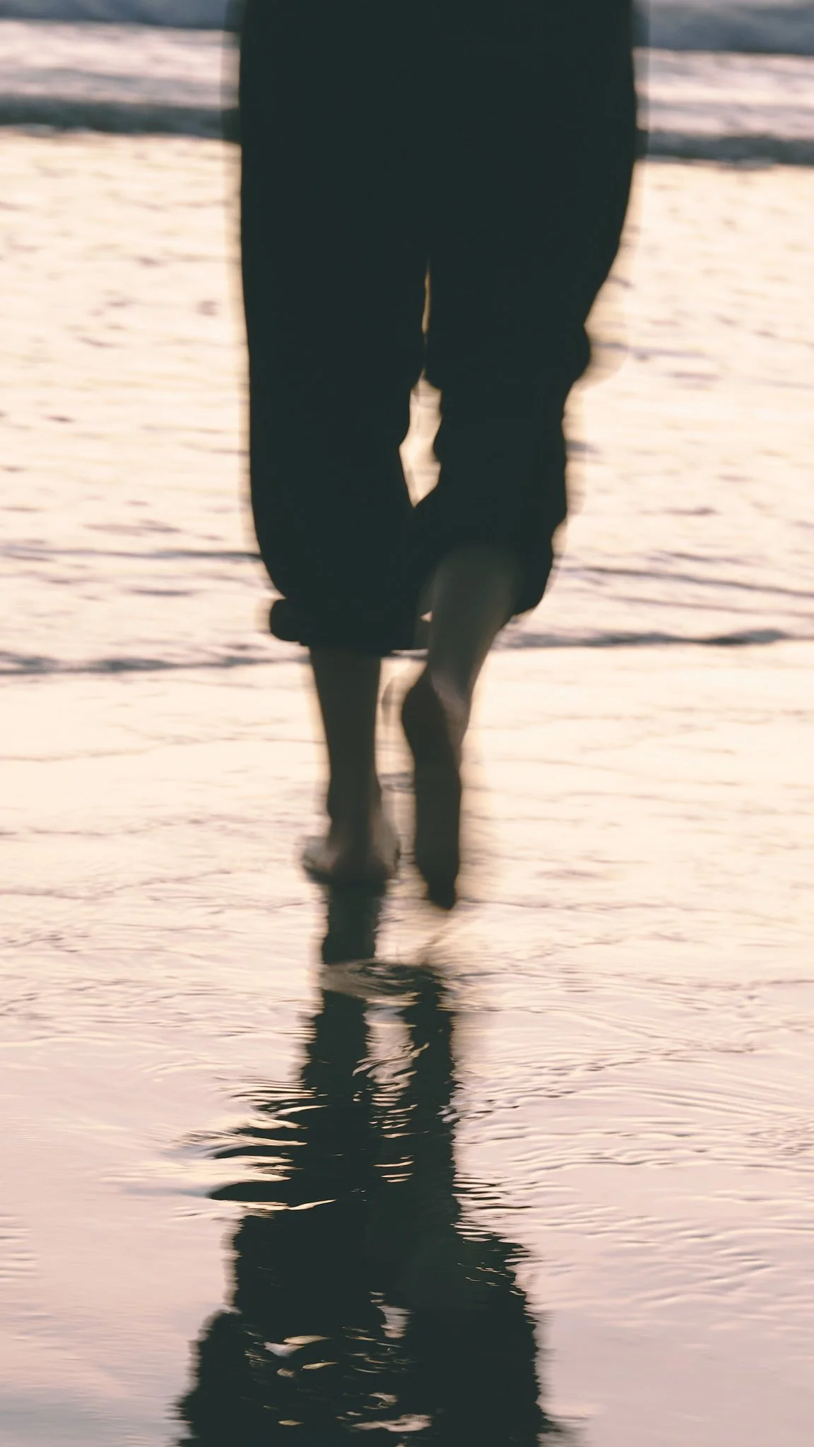A person walking barefoot on the beach at sunset, reflected in the wet sand.
