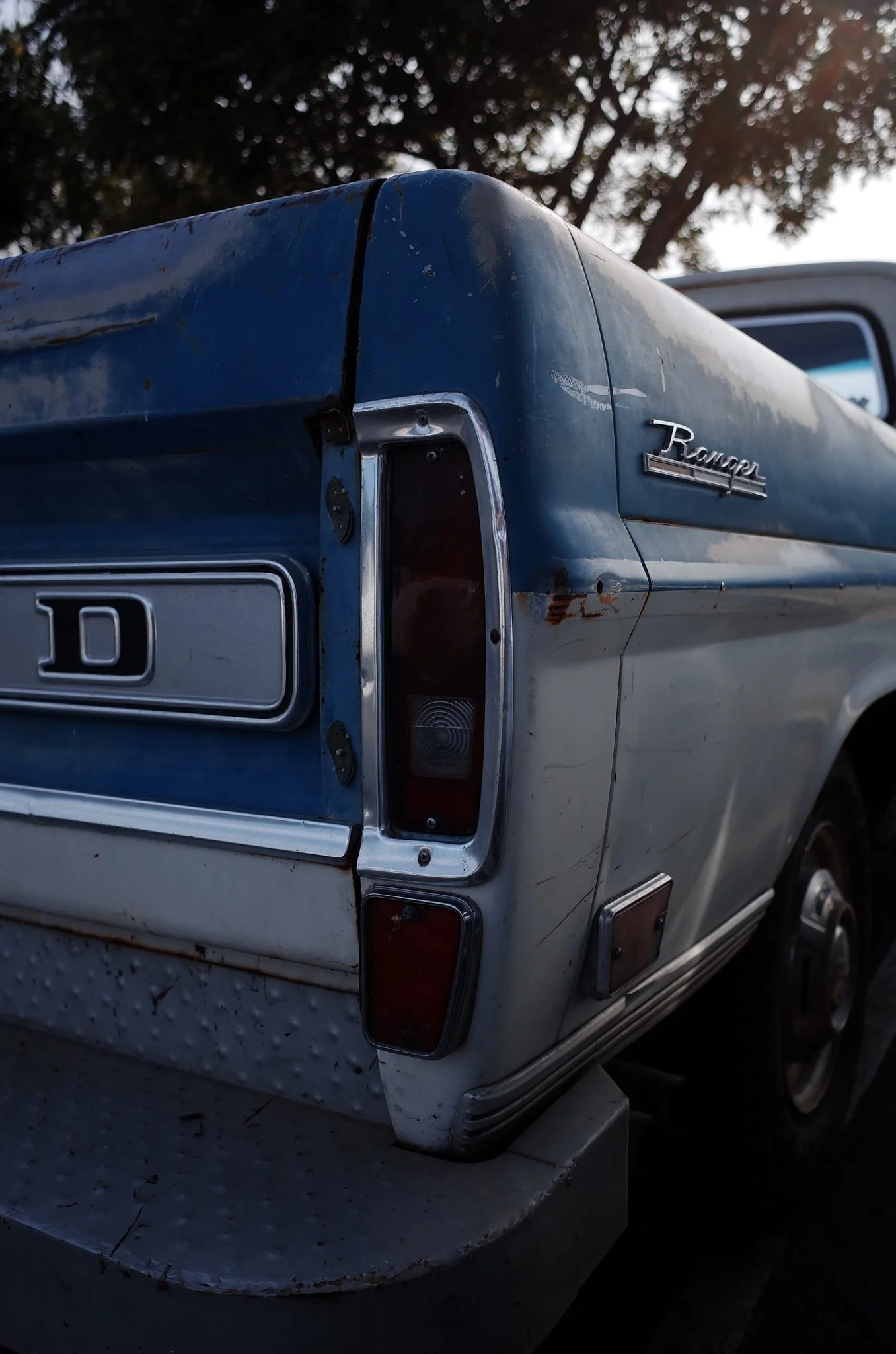 Close-up of the rear of a blue vintage Ford Ranchero pickup truck, showing rust, scratches, and the 'Ranchero' badge, with a tree and sky in the background.