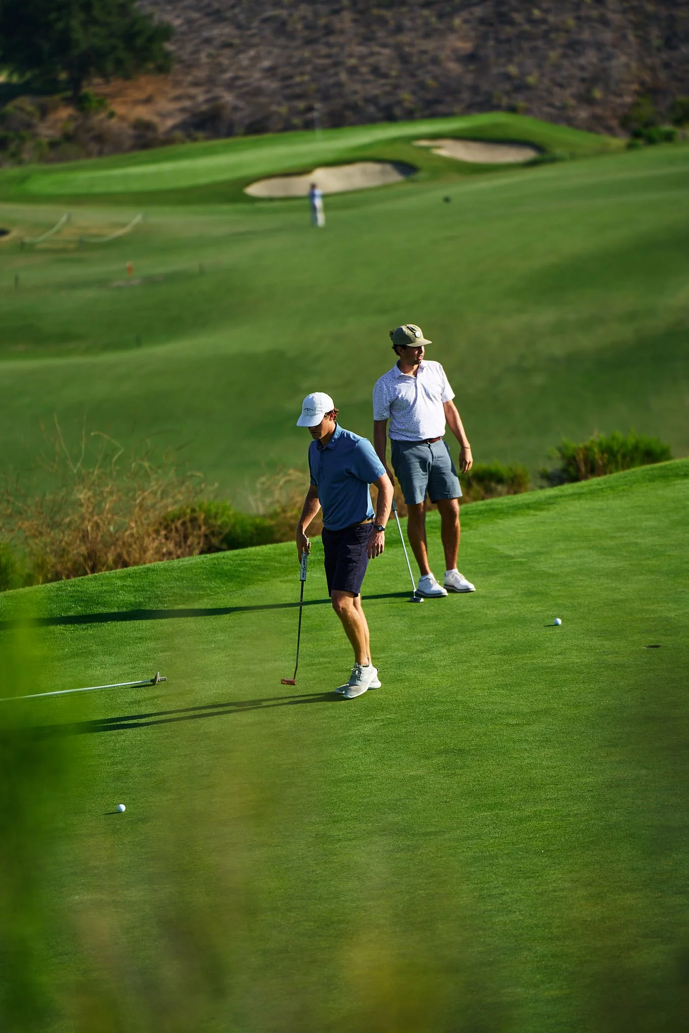 Two men playing golf on a lush green golf course with sand bunkers and hills in the background; one man is preparing to putt while the other observes.