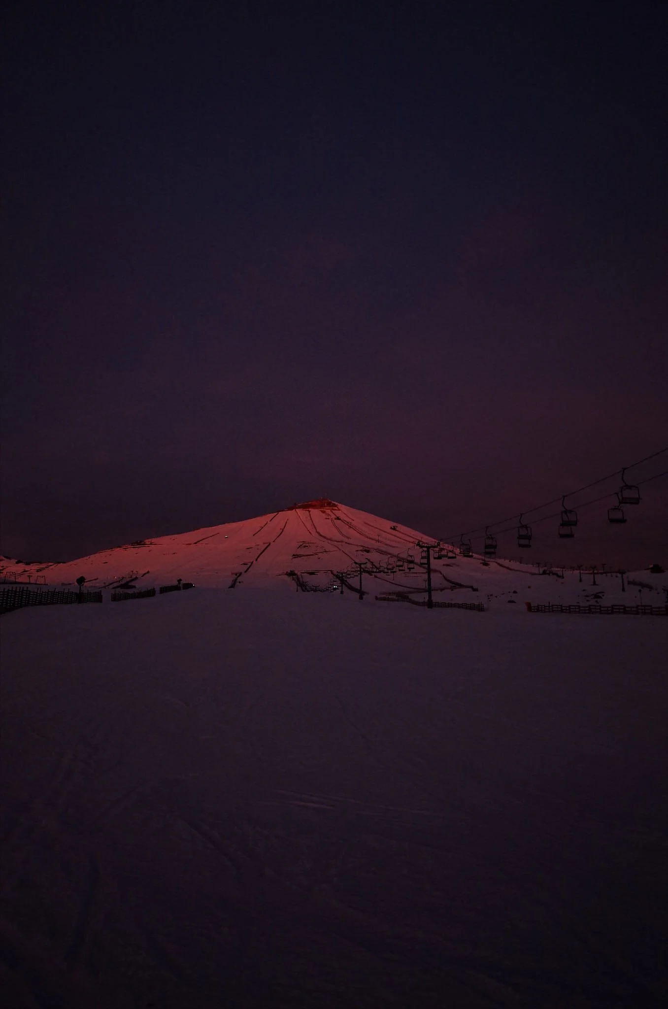 Snow-covered ski slopes at night with a distant mountain illuminated in pinkish light and ski lift chairs visible against a dark sky.