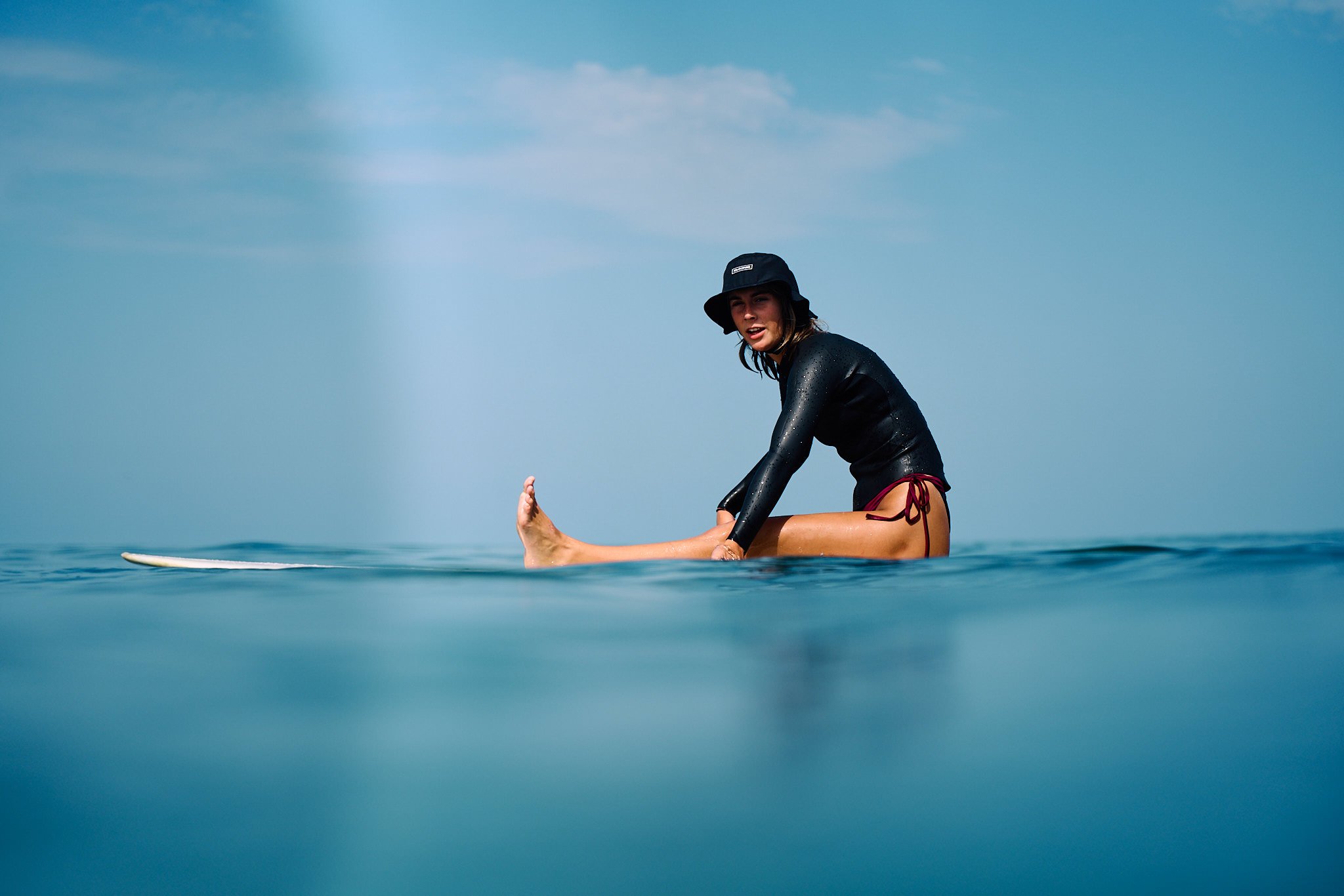 Young woman in black wetsuit and bucket hat practicing yoga on a surfboard in the ocean under a partly cloudy sky.