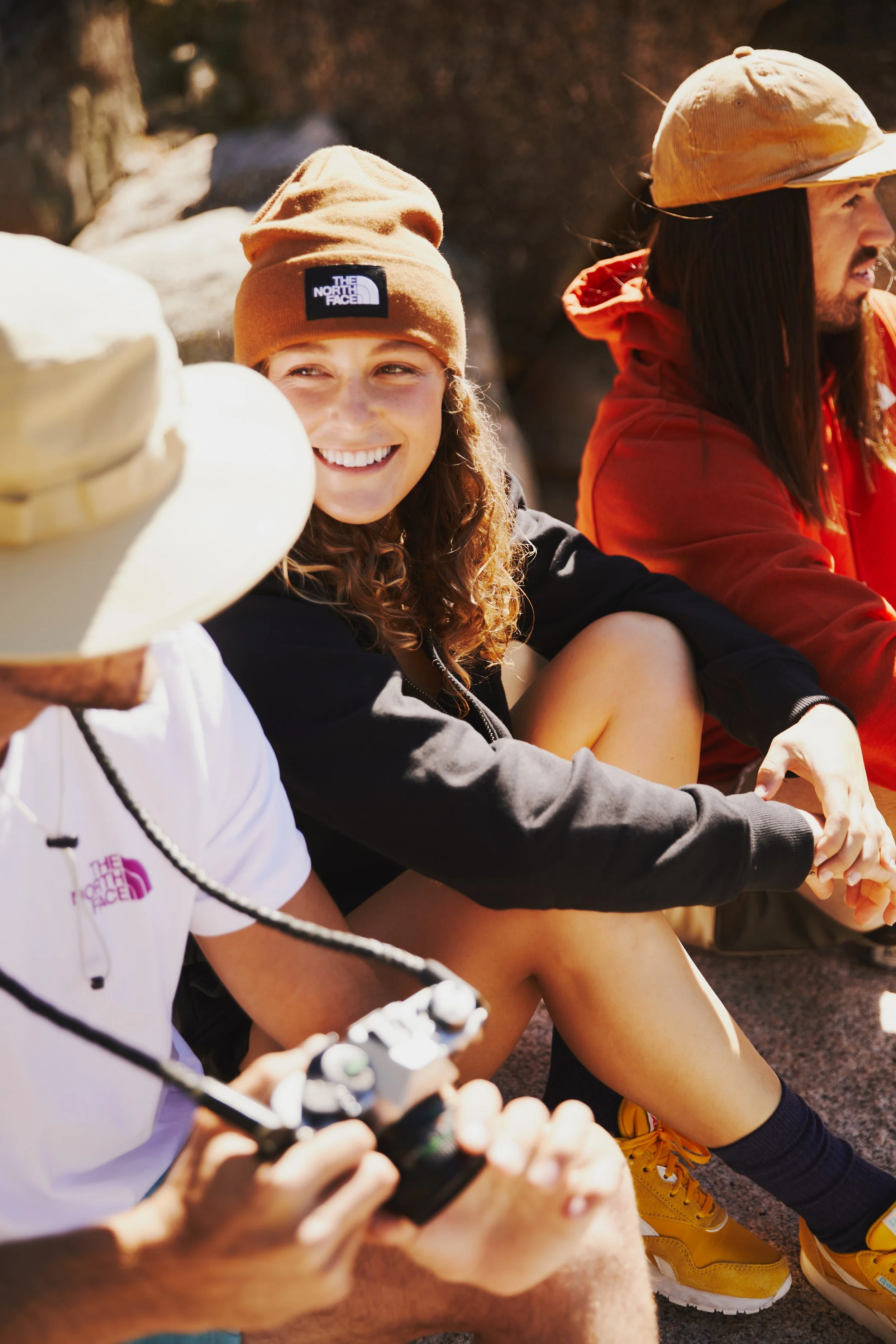 Three people sitting outdoors: a young woman in a brown beanie with The North Face logo, a man in a tan cap and red hoodie, and another person in a white shirt with a wide-brim hat. They are smiling and looking in different directions.