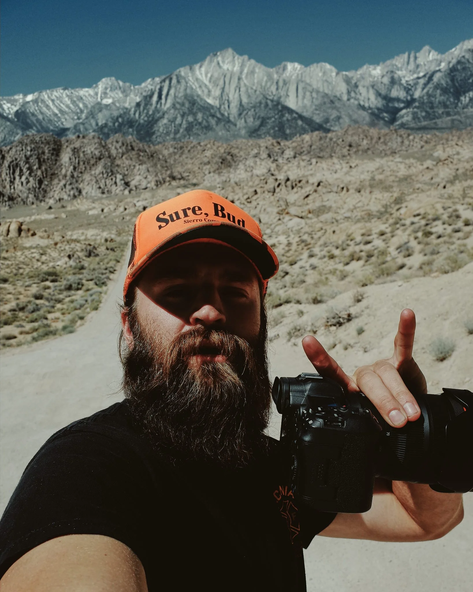 A man with dark hair and a beard wearing an orange cap that says "Sure, Bud" and a black shirt, taking a selfie in a rocky, mountainous landscape with snow-capped peaks in the background, holding a camera in one hand and making a peace sign with the 