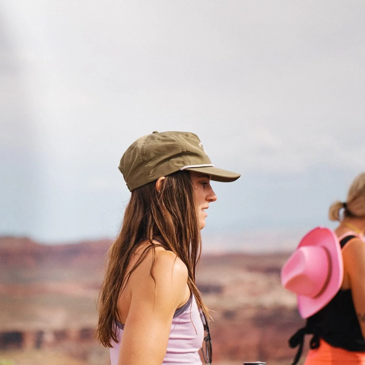 Side profile of a woman with long brown hair wearing a tan cap and a sleeveless top, outdoors with a blurred desert landscape in the background.