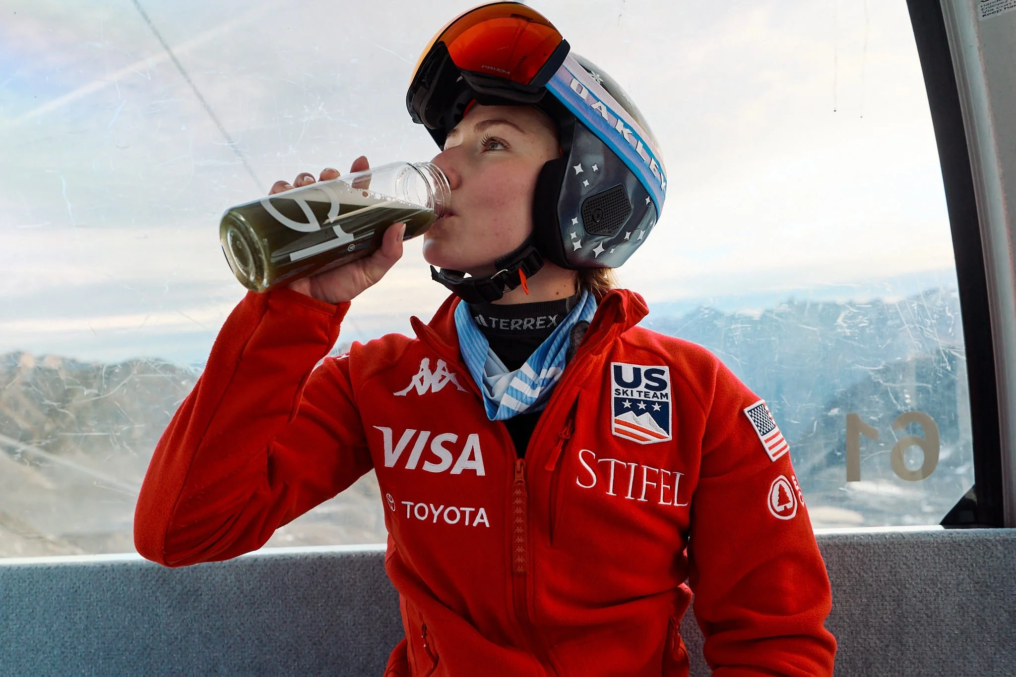 A person wearing a blue ski helmet and red jacket with sponsor logos, drinking from a glass bottle while observing the landscape from inside a ski lift cabin.
