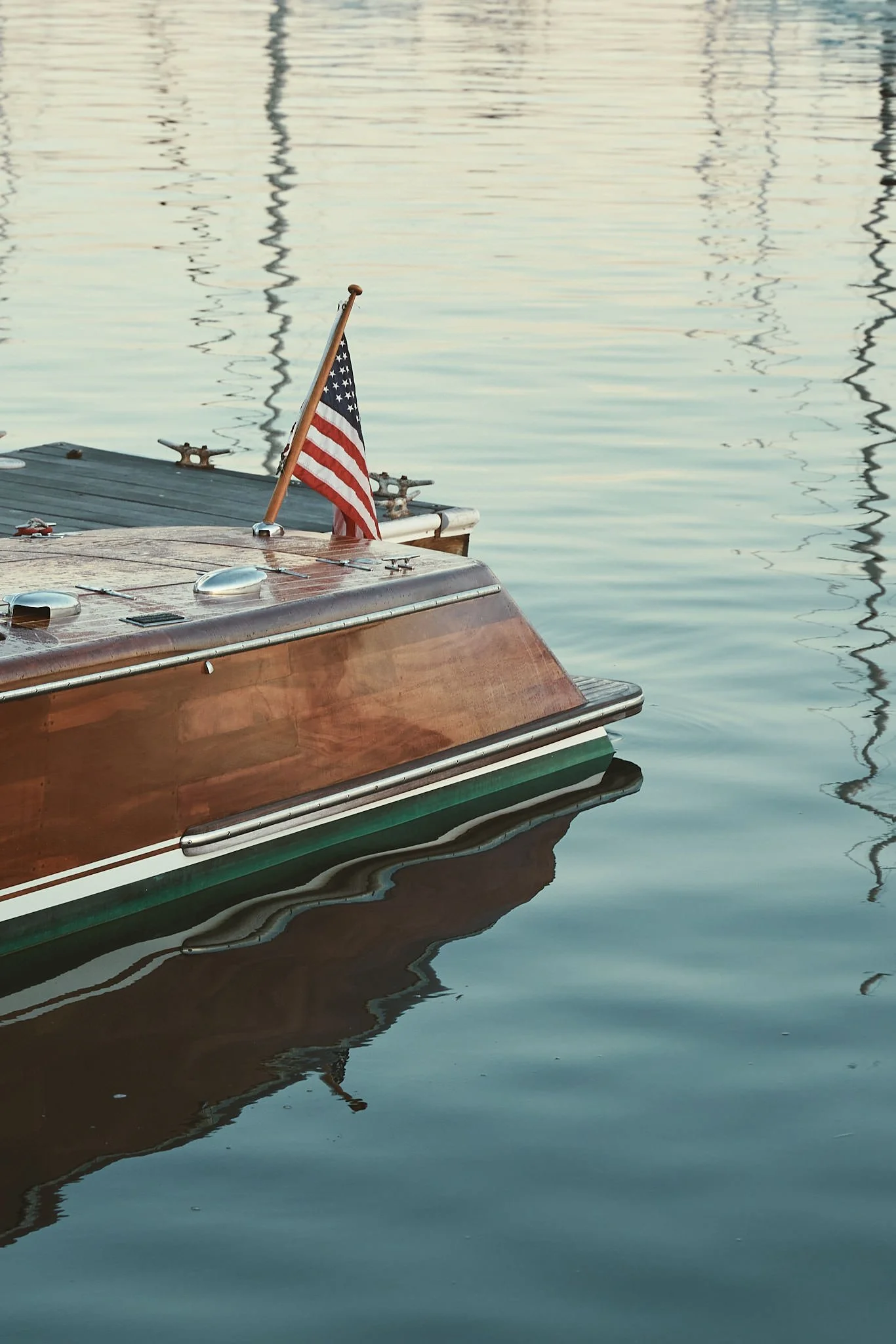 A boat with a small American flag attached to a wooden pole, floating on calm water with reflections of masts visible in the background.
