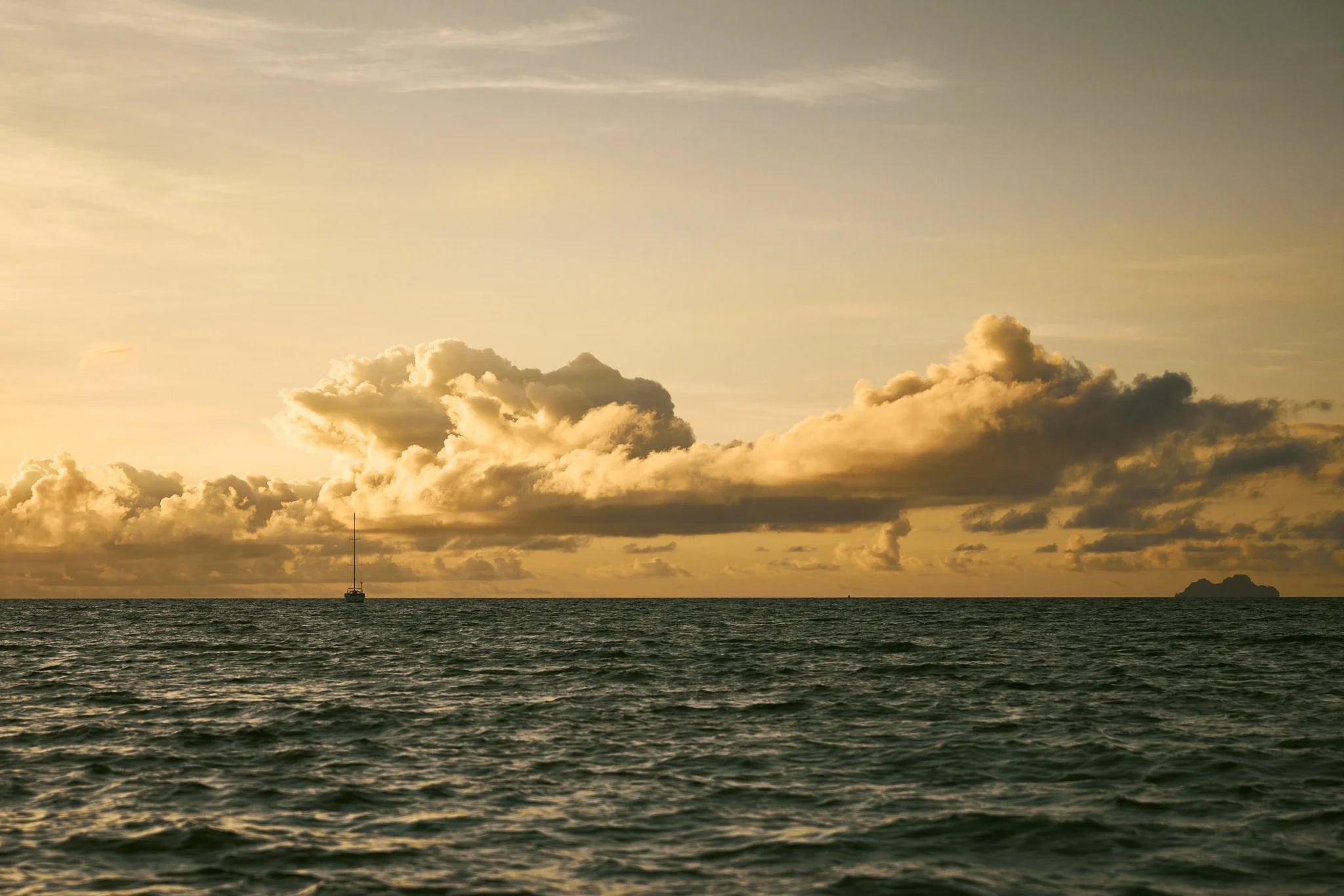 Sunset over the ocean with a sailboat and a small island in the distance.