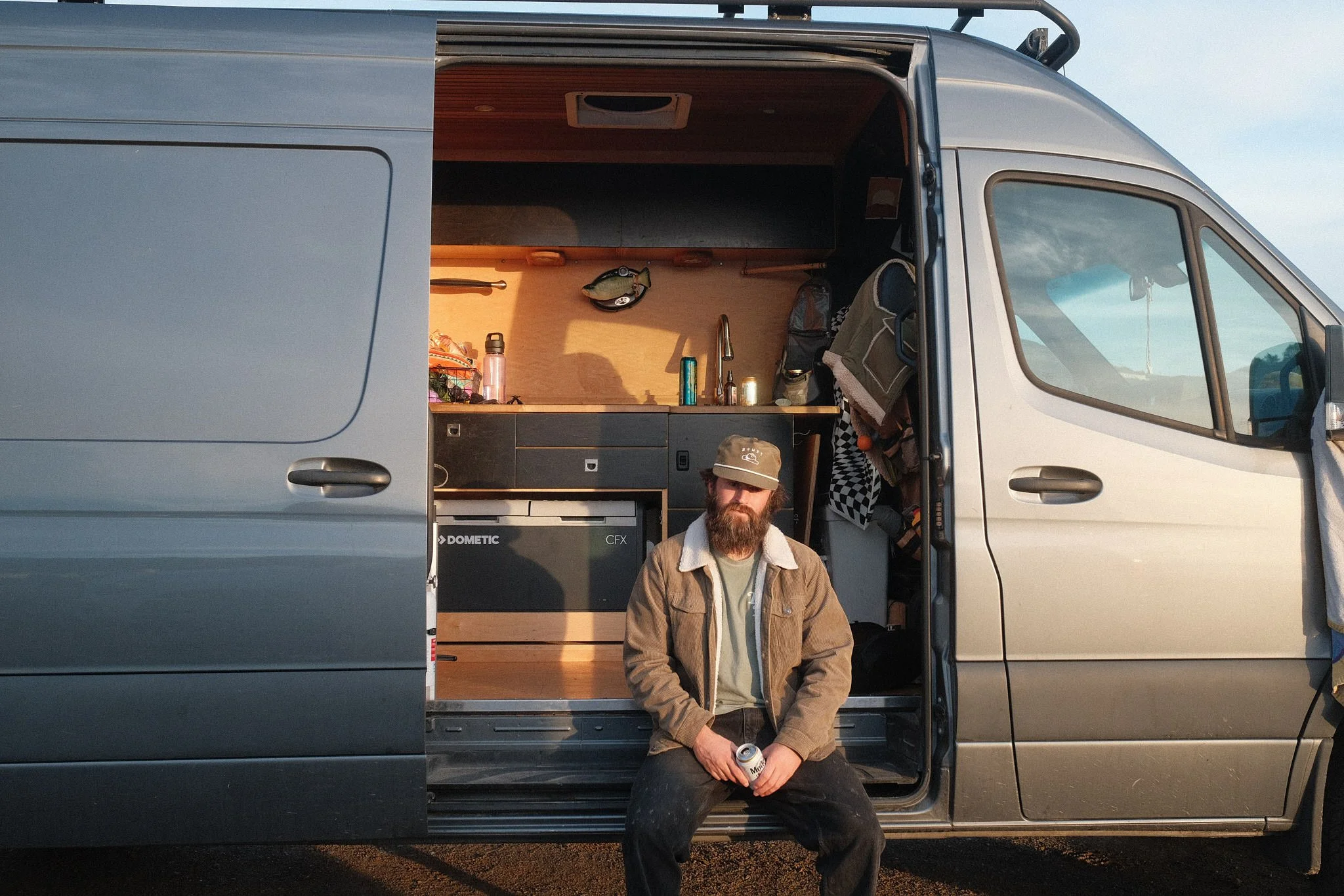 A man with a beard and a cap standing in front of a silver camper van, holding a can of beer. The van has an open side door revealing a compact kitchen area inside with a sink, cabinets, and various items.
