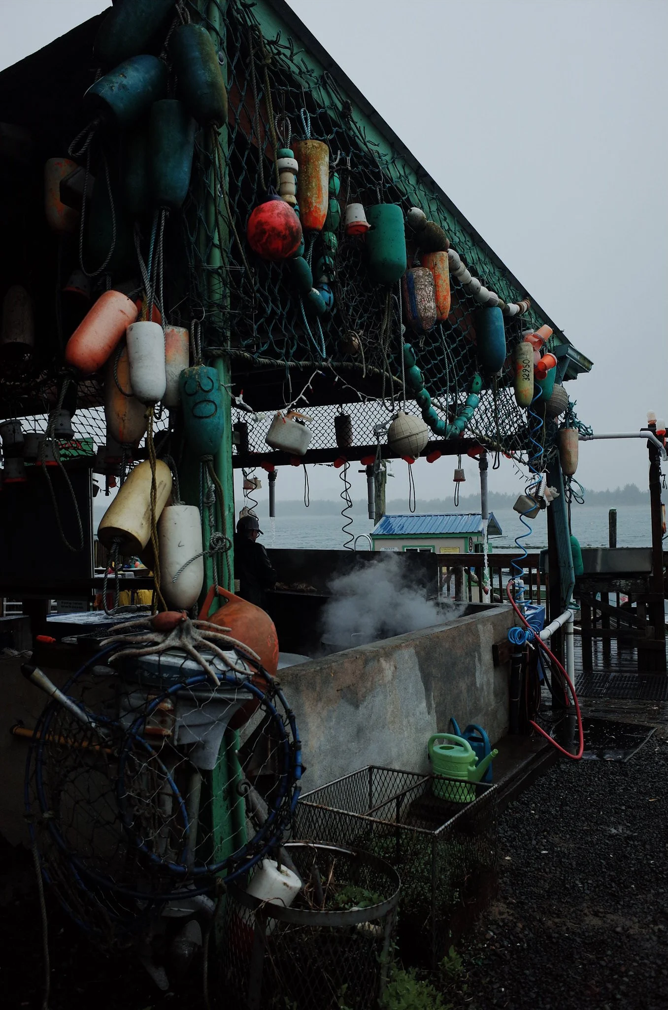 Fishing boat dock with floating buoys, fishing gear, and a person wearing a hat, against a cloudy sky and water background.