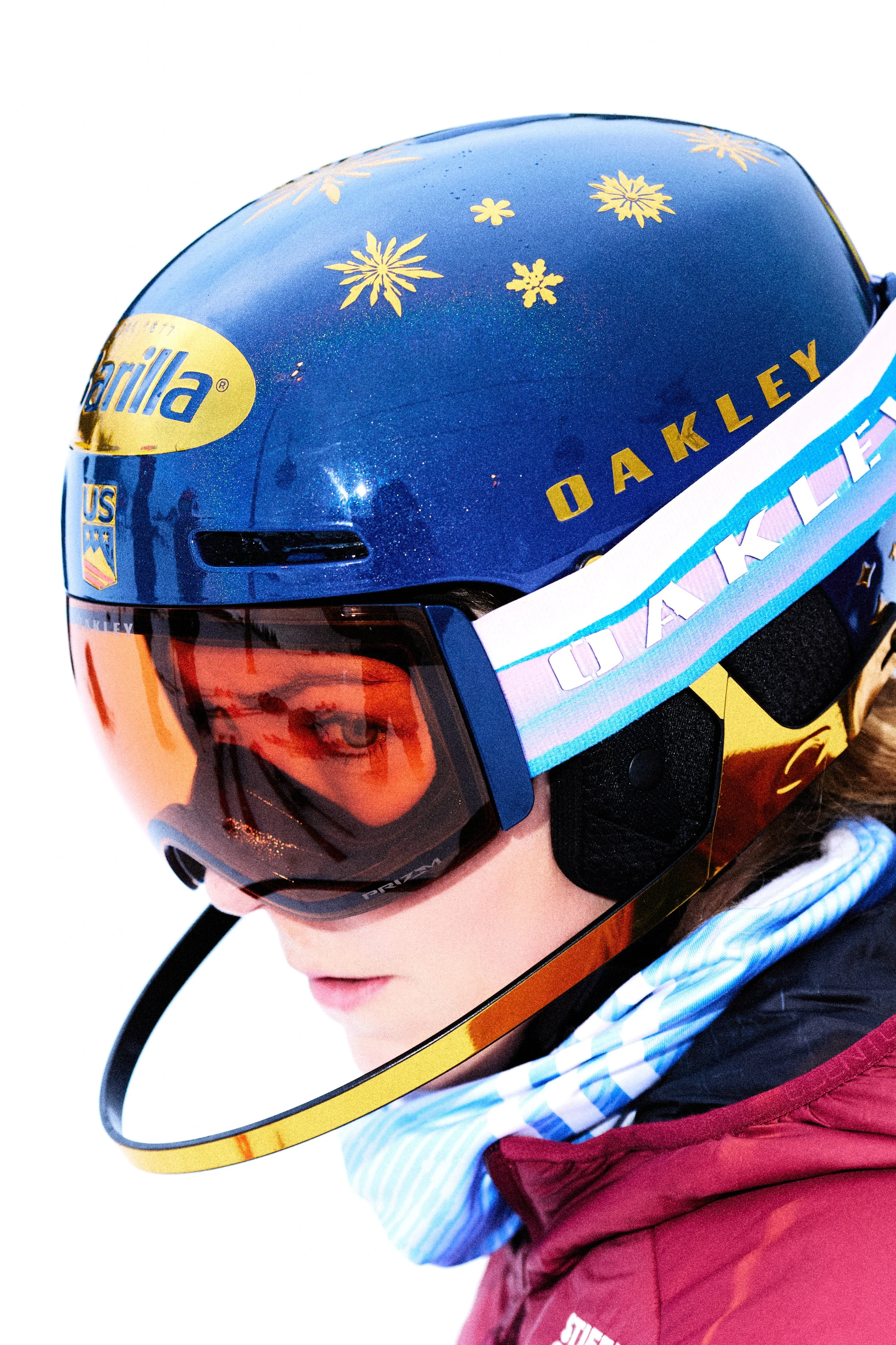 Close-up of a young person wearing an oakley helmet with a blue and yellow design and gold snowflake patterns, wearing ski goggles, a striped blue and white shirt, and a maroon winter jacket.