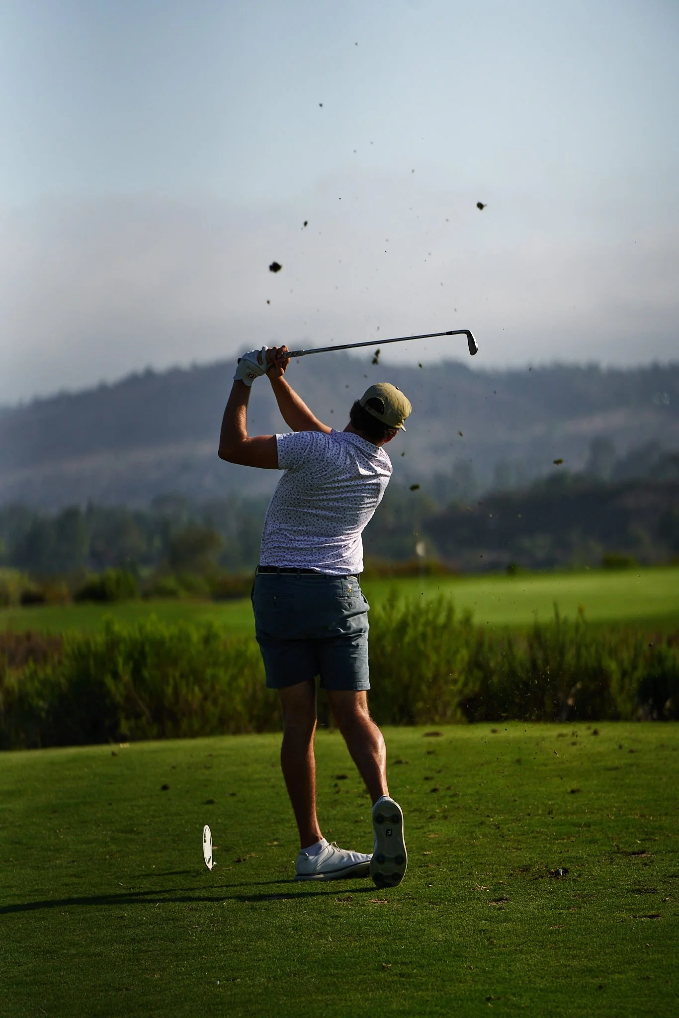 A male golfer swings a golf club on a golf course, with dirt and grass flying up as he hits the ball, against a background of trees and distant hills under a partly cloudy sky.