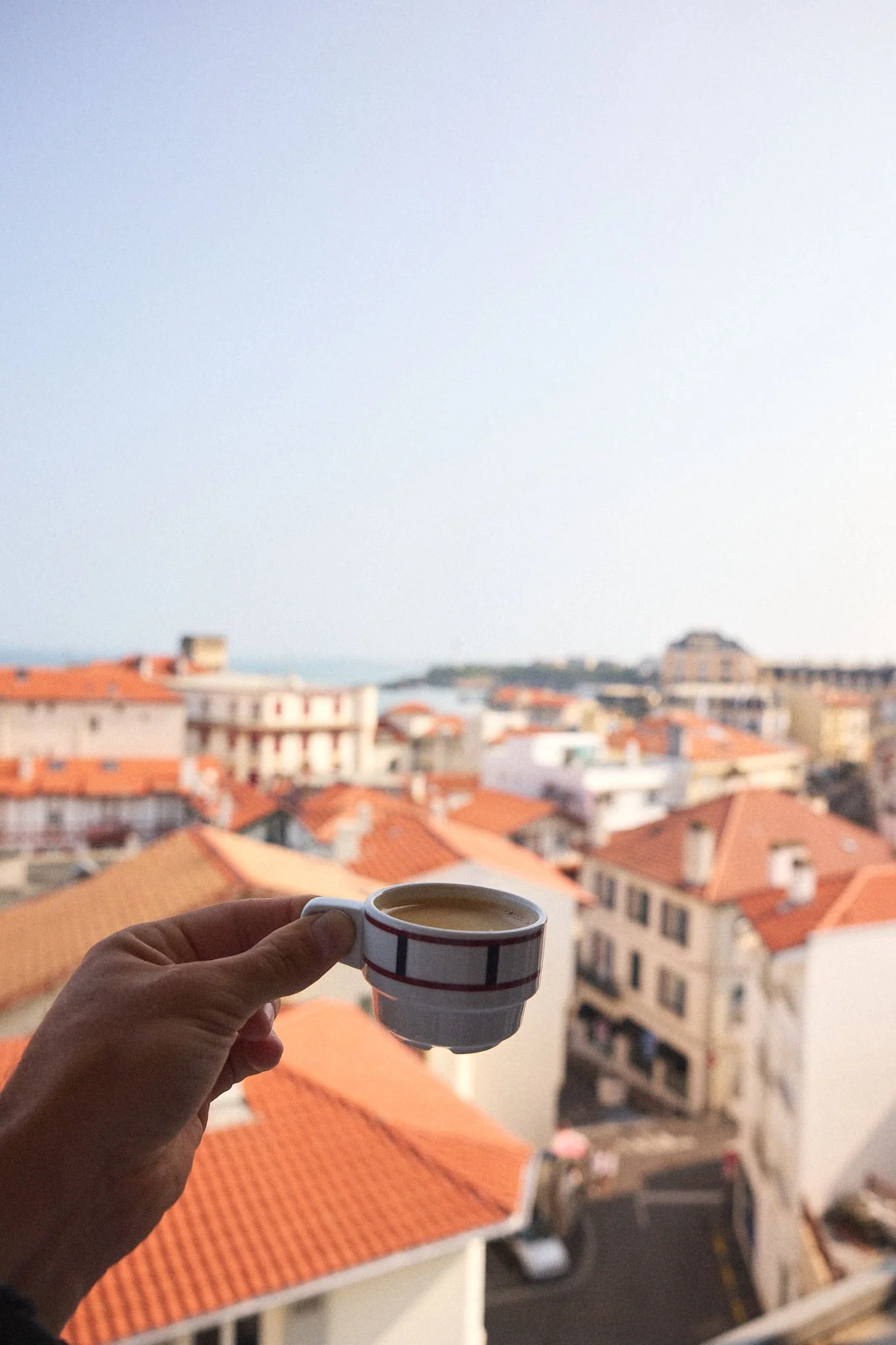 A person holding a small striped coffee cup with a cityscape of buildings with orange rooftops and a distant body of water in the background