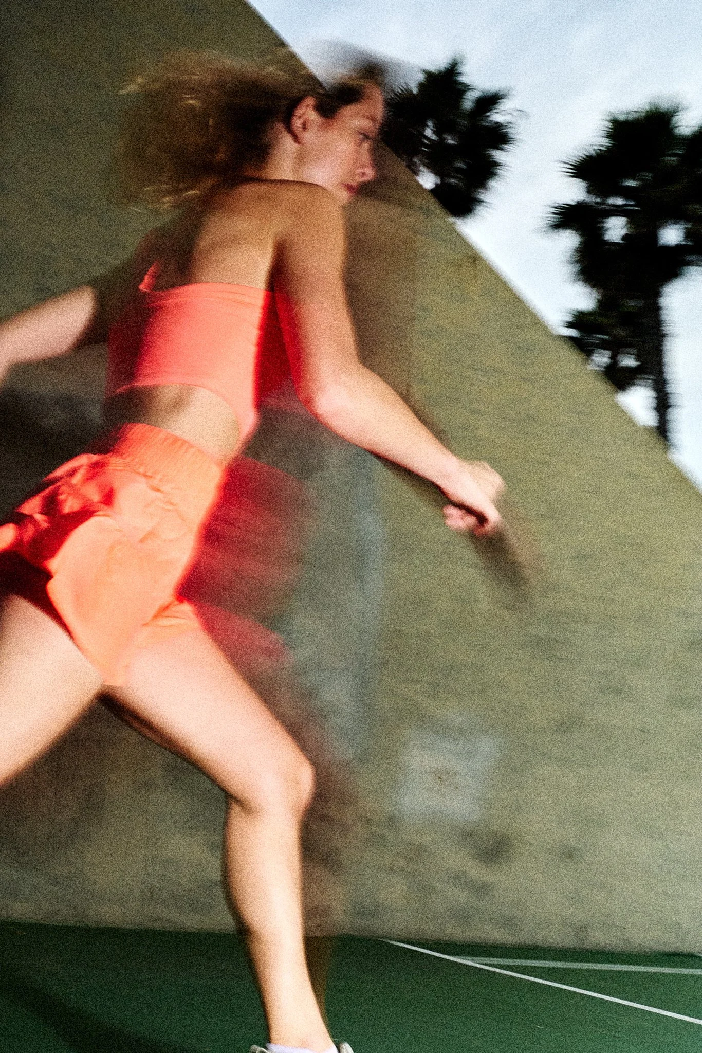 A woman with curly hair wearing a coral sports bra and matching shorts lying on a tennis court, with palm trees in the background, taken during dusk or dawn.