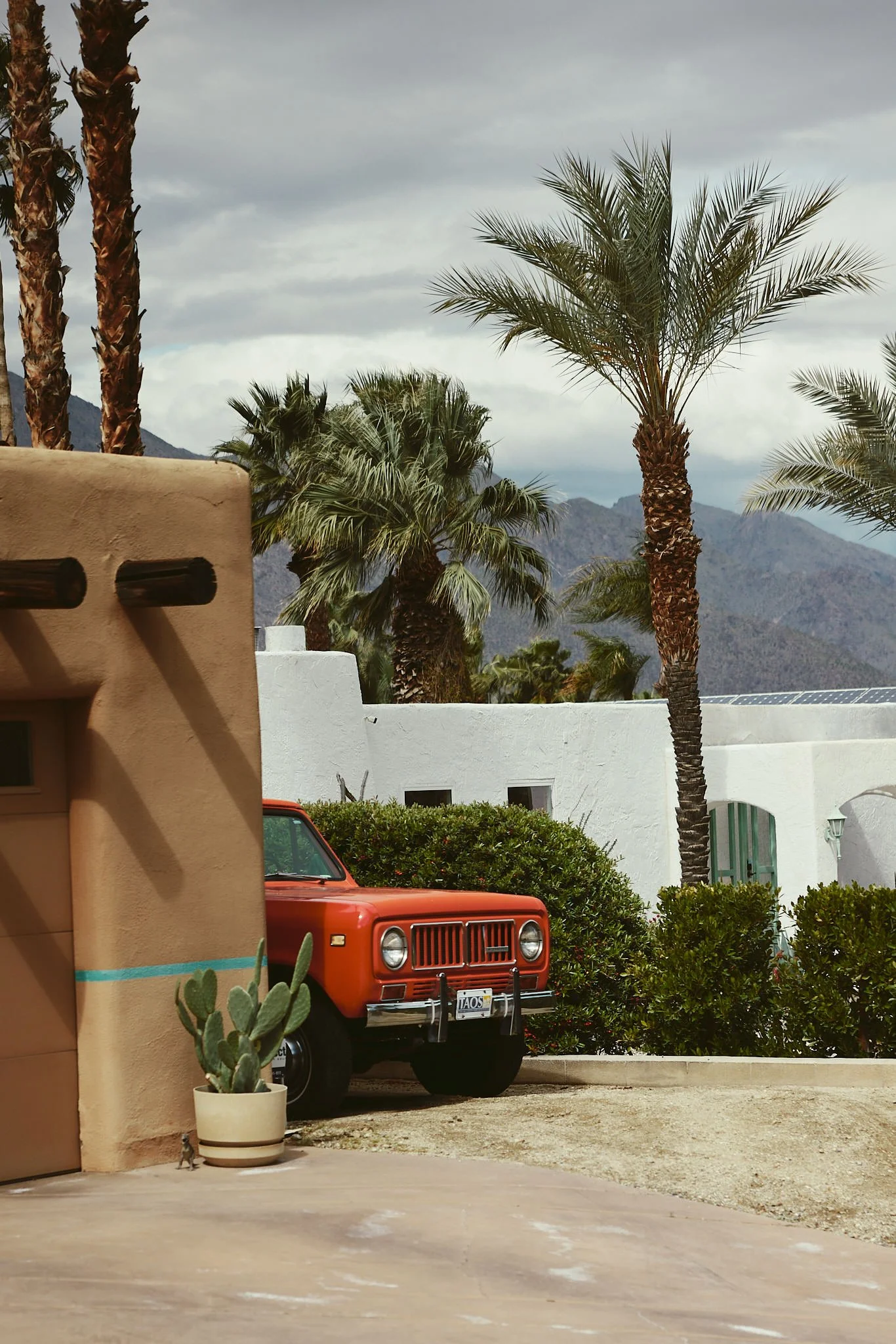 View of a neighborhood with palm trees, mountains in the background, and a red vintage Jeep parked behind a white wall, partially visible.