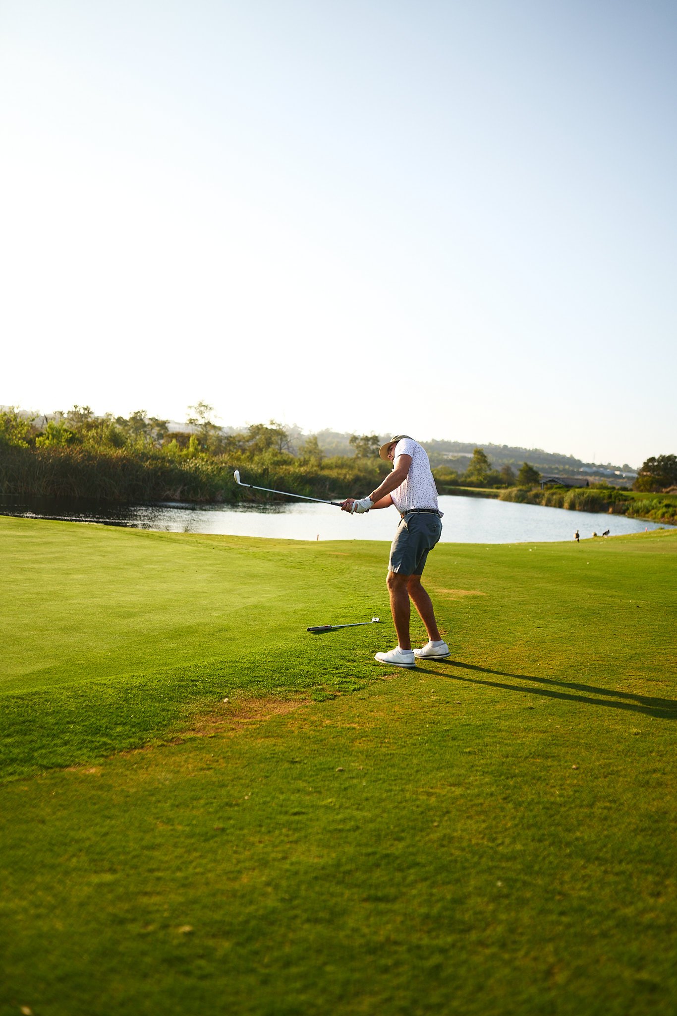 A man playing golf on a lush green course near a body of water under a clear sky.