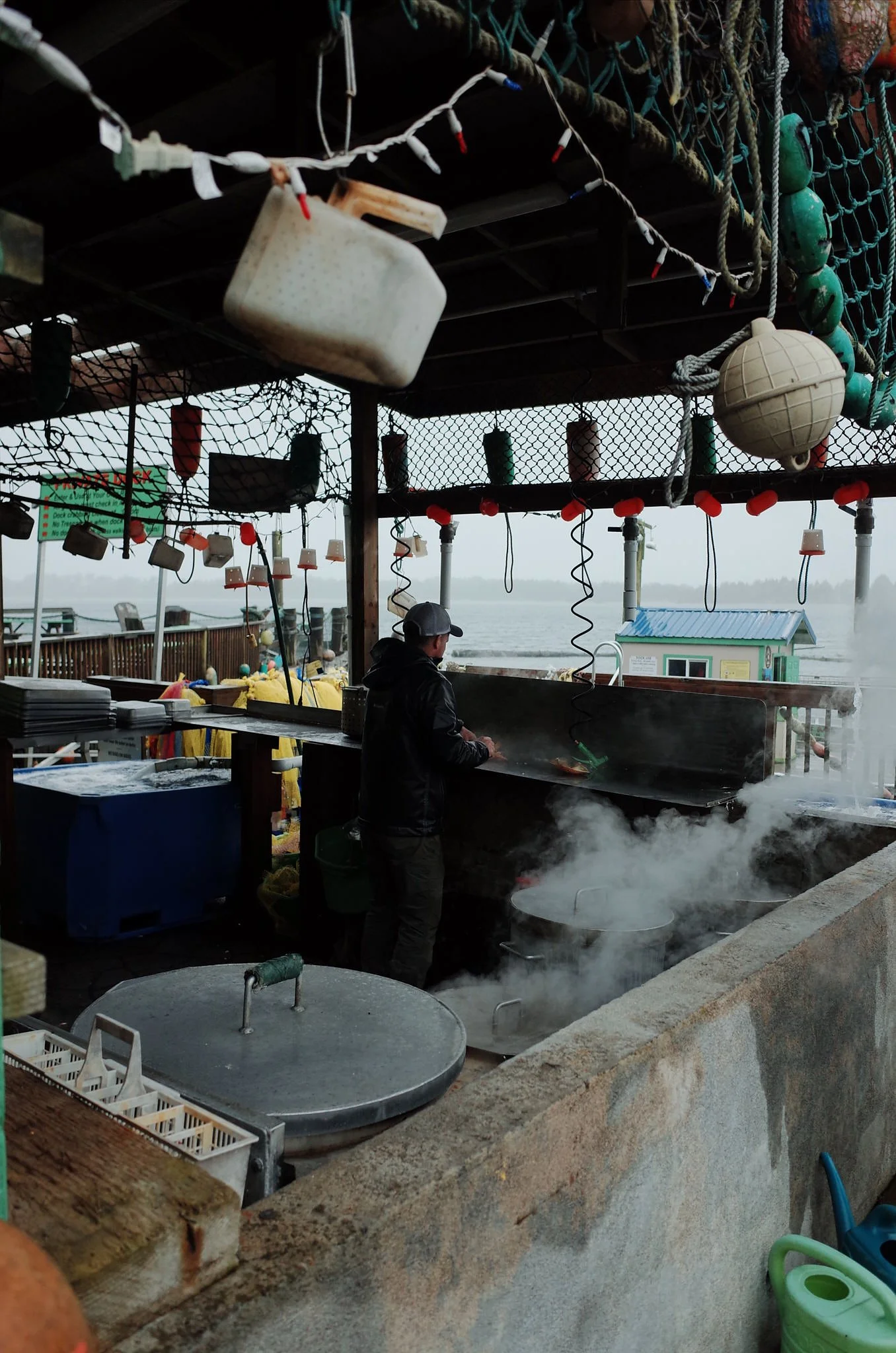 Fish cleaning station with a man cooking fish over a grill, surrounded by fishing buoys, ropes, and nets at a dock near the water.