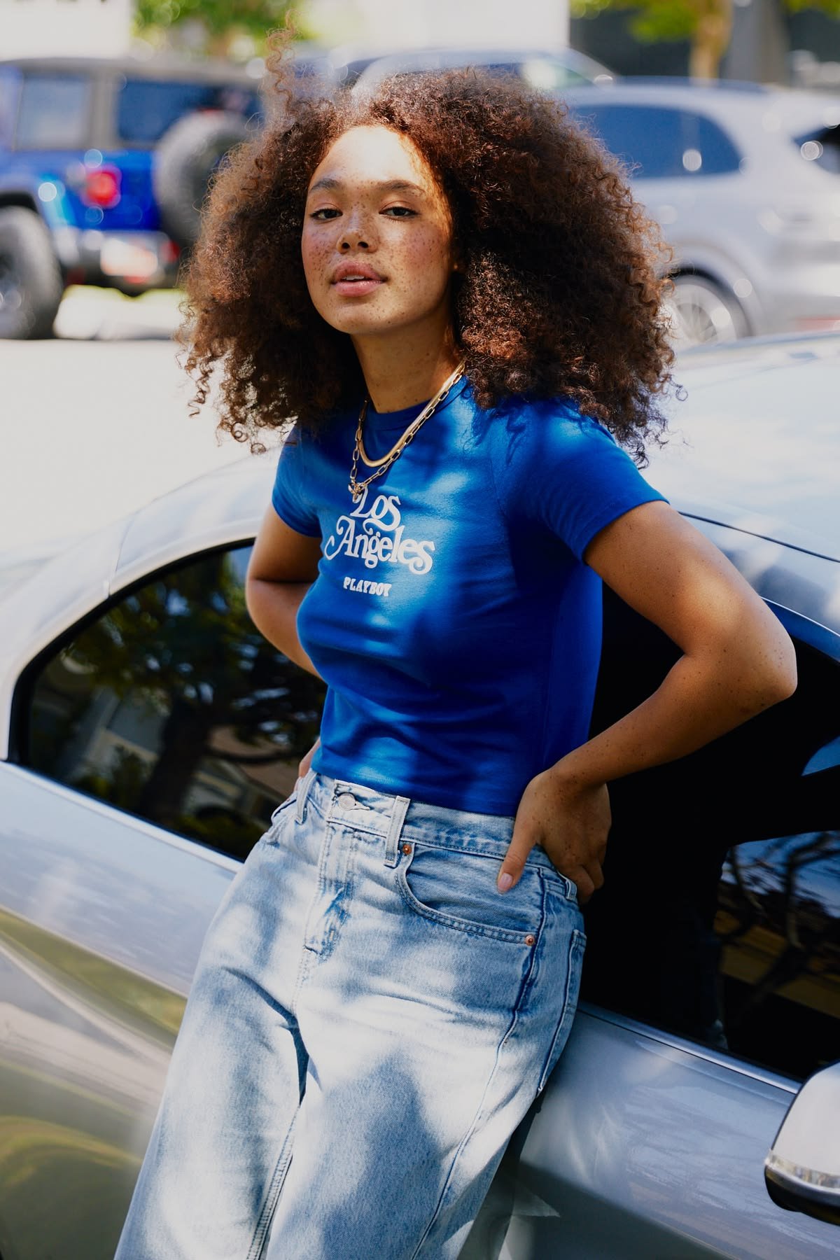 A young woman with curly hair and freckles wearing a blue 'Los Angeles' t-shirt and light denim jeans, leaning against a white car in a parking lot.