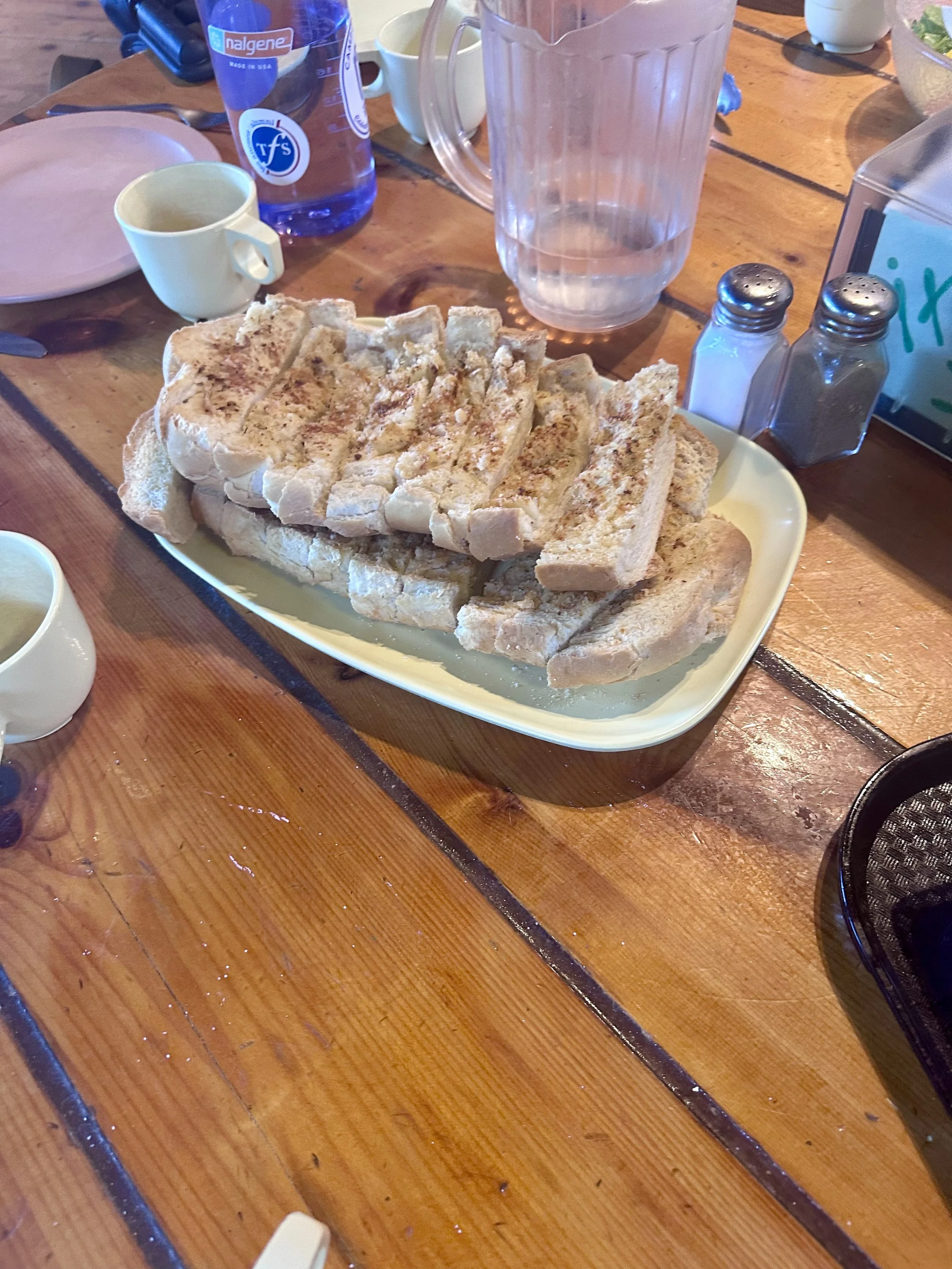 A basket of sliced white bread on a yellow plate on a wooden table, with salt and pepper shakers, a pitcher of water, and various condiments and utensils nearby.