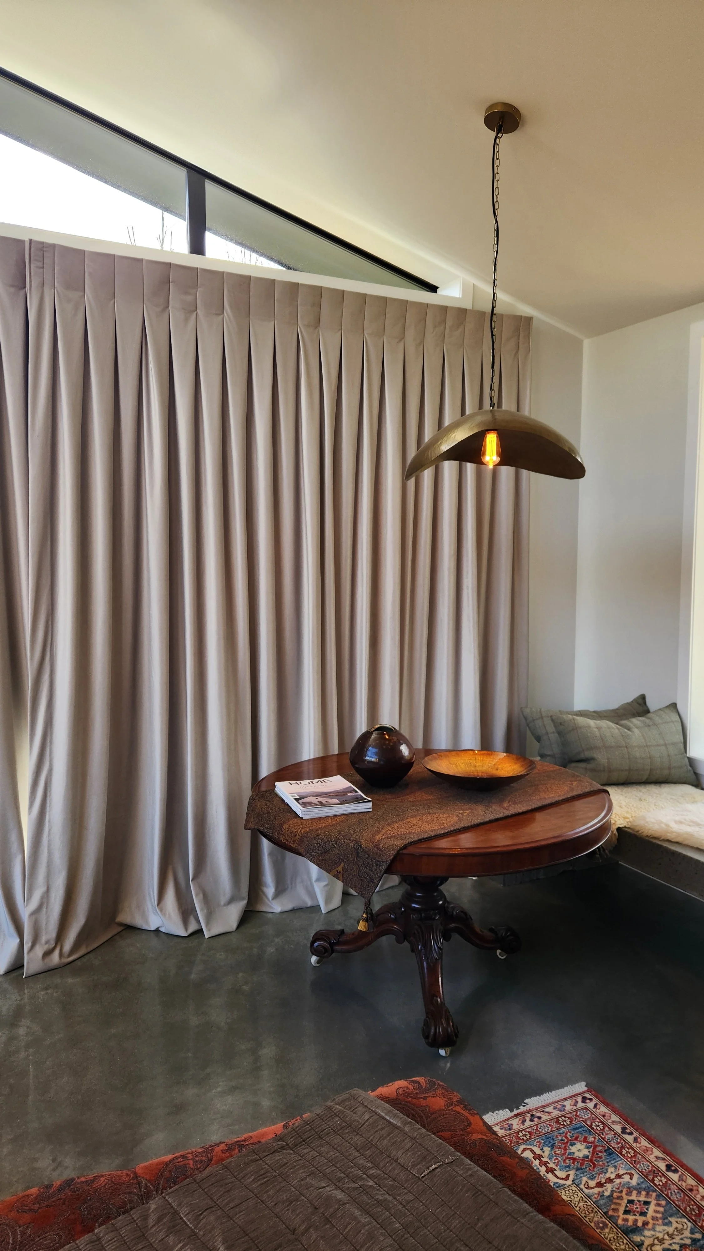 Interior of a room with beige curtains, a wooden table with a decorative cloth, a dark ceramic bowl, a magazine, and a pendant light hanging from the ceiling. Part of a couch with patterned cushions and a rug are visible.