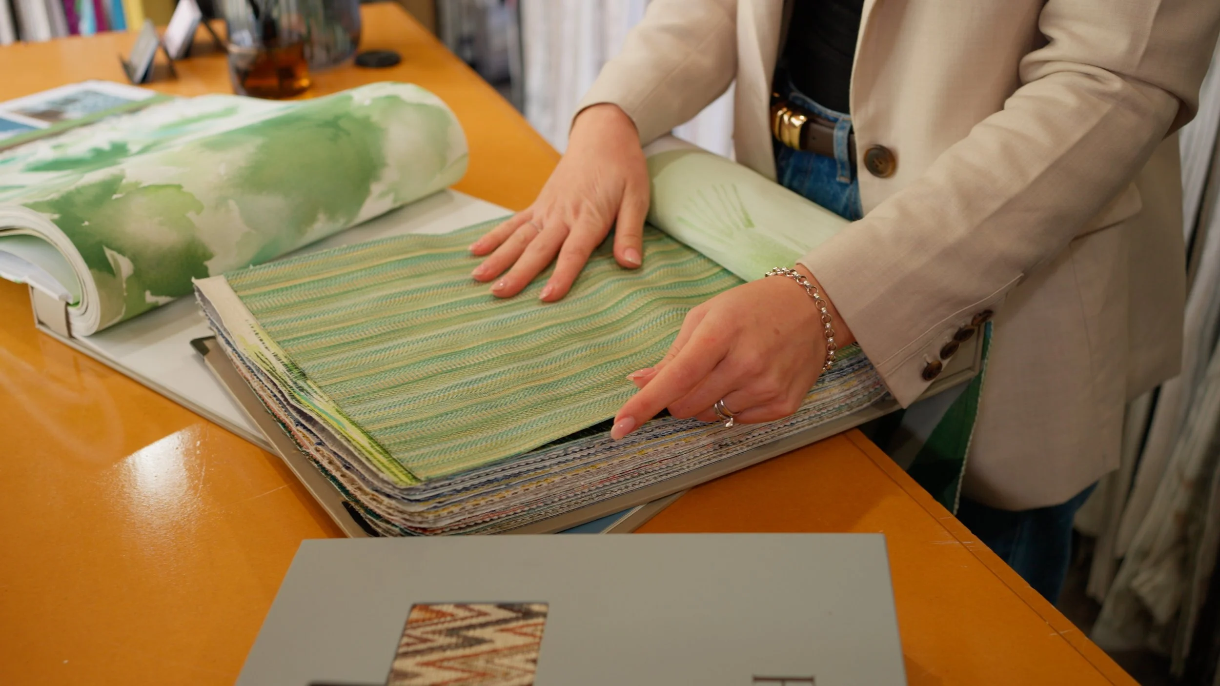 Person flipping through fabric swatches or samples on a table, with various fabric pieces in shades of green and patterns. The person is wearing a beige blazer, a black top, and blue jeans, with jewelry on their wrist and finger.
