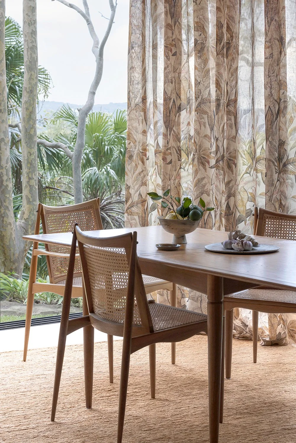 A wooden dining table with four chairs, a bowl with green leaves, and seashells, inside a room with patterned curtains, overlooking outdoor greenery with trees.