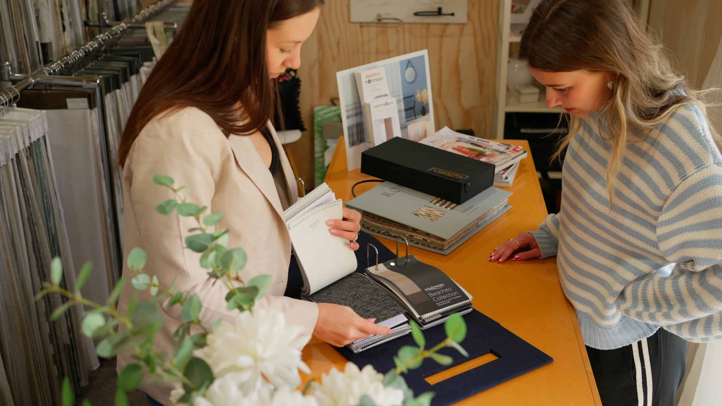 Two women are in a retail store, examining fabric samples on a table with brochures and catalogs. One woman is holding a fabric sample book, and the other is leaning over the table, looking at the samples.