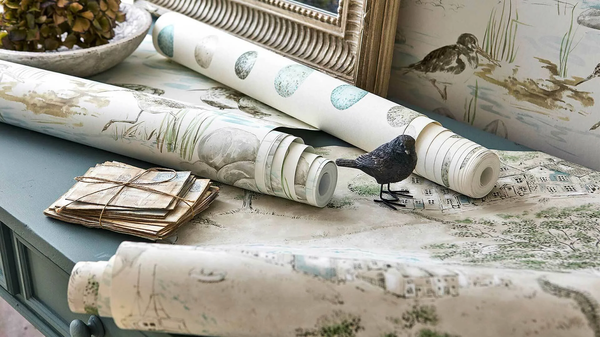 A tabletop with rolled-up water-themed wallpaper, a small stack of vintage postcards tied with string, a framed mirror, and a bird figurine in front of a nature-inspired wallpaper background.