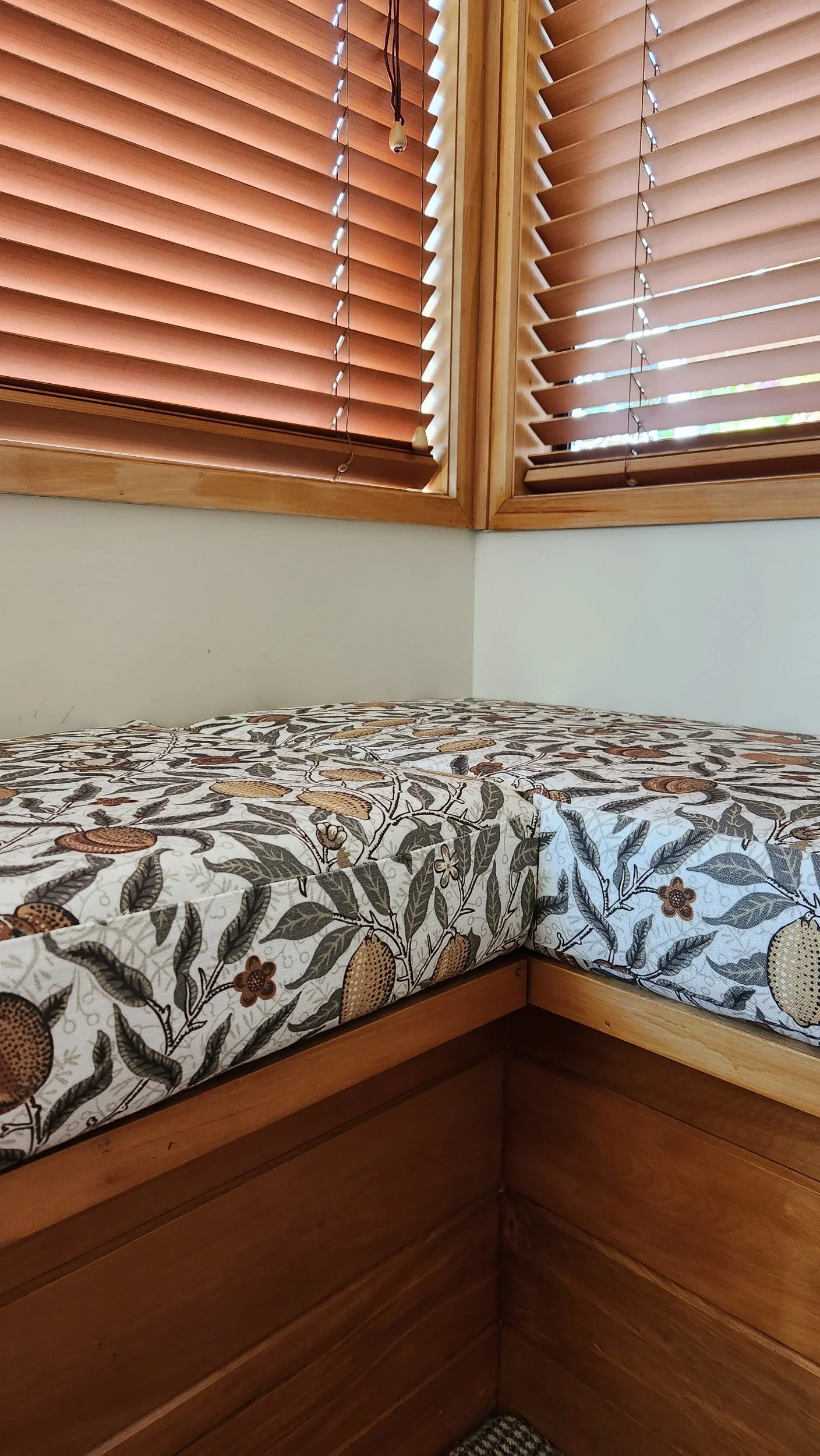 Corner of a bedroom with wooden window blinds and a floral-patterned mattress on a wooden bed frame.