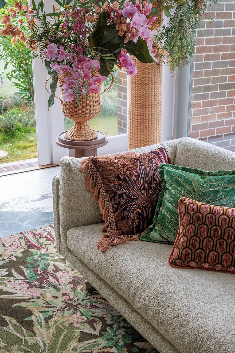 Living room with a beige sofa adorned with decorative pillows in green, pink, and brown patterns. Behind the sofa is a large floral arrangement in a woven basket, placed on a small round table. The arrangement includes pink orchids and various greenery. The room has a large window at the back, with a view of a garden, and a brick wall is visible on the right. A floral-patterned rug covers the floor.