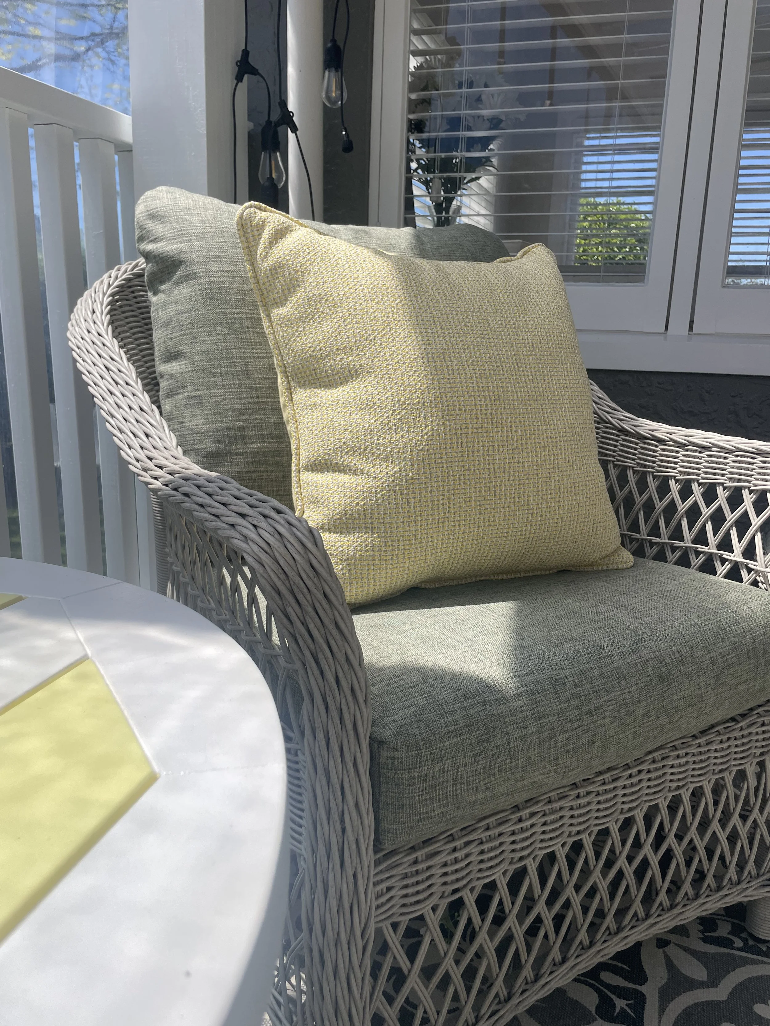 A wicker outdoor sofa with beige cushions on a porch or patio, next to a window with white blinds and a string of outdoor string lights.