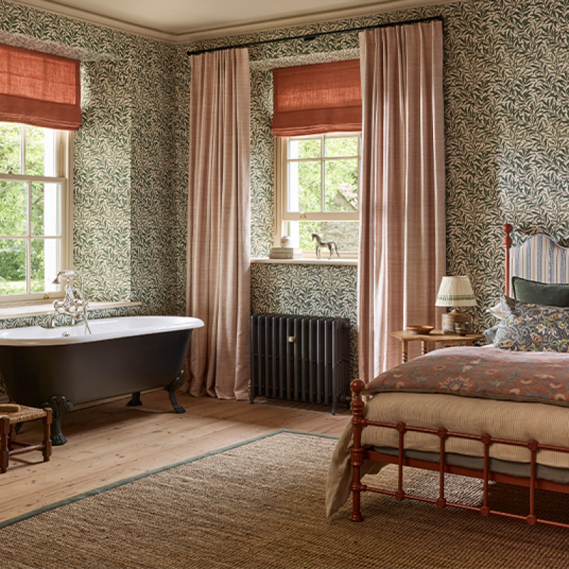 A bedroom with vintage decor, featuring a copper bed frame, patterned wallpaper, a bedside table with a lamp, a window with pink curtains, and a freestanding bathtub beside the window.