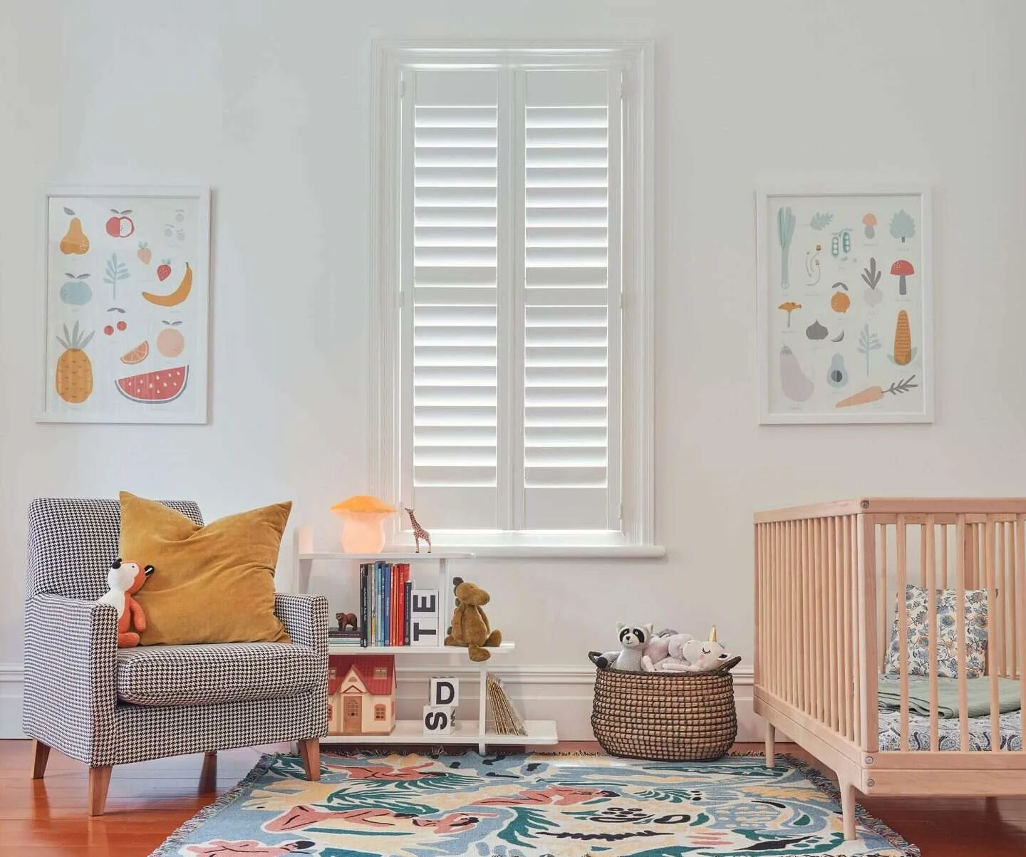 A nursery room with white walls, a window with white shutters, framed animal and fruit artwork, a black and white checkered armchair with yellow pillows and plush toys, a white bookshelf with books, a plush bear, a basket with plush toys, a wooden crib, a colorful rug, and decorative toys and art.