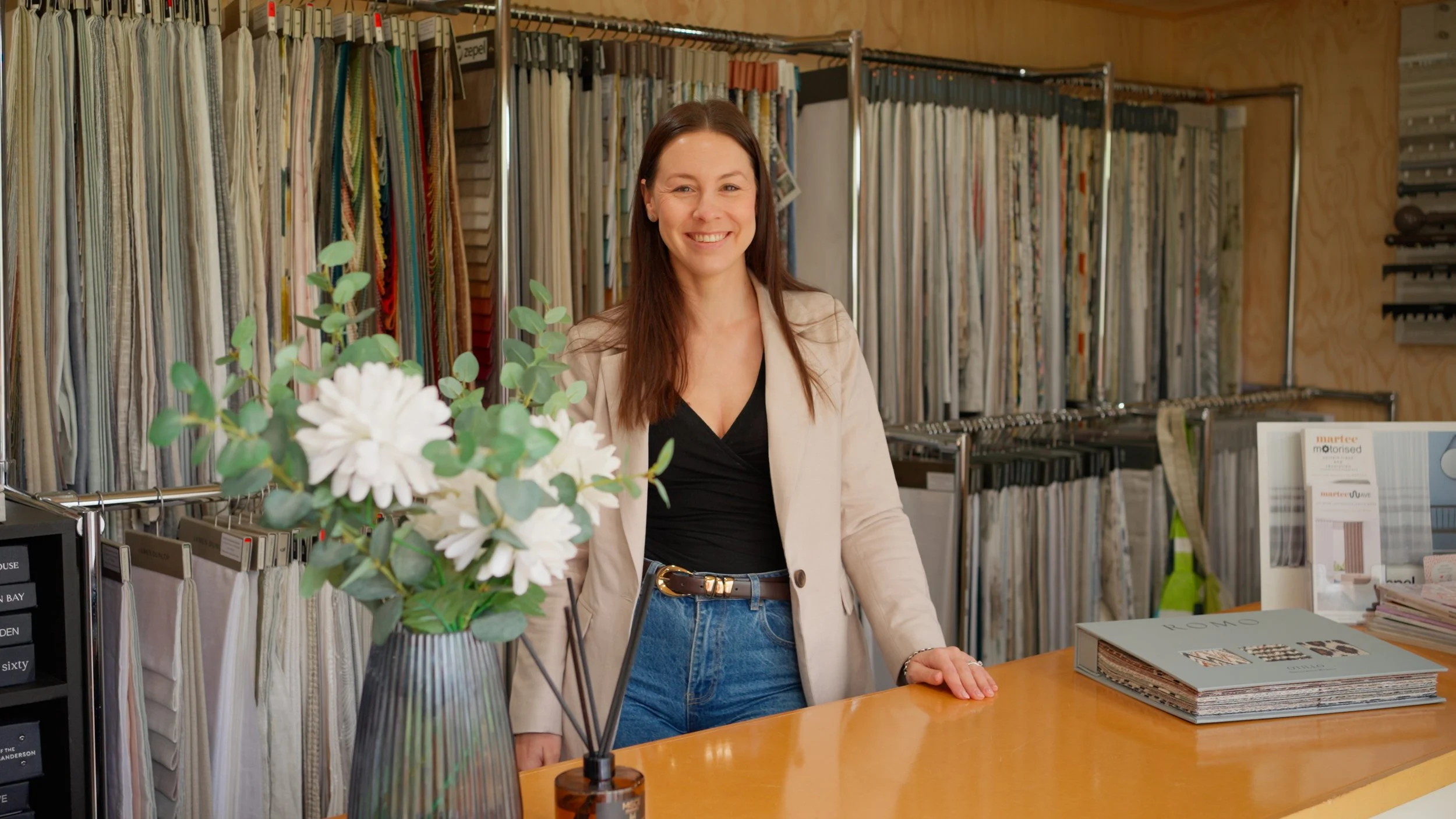A woman with long brown hair smiling behind a counter in a fabric store, with fabric swatches hanging on racks behind her and a vase of white flowers on the counter.