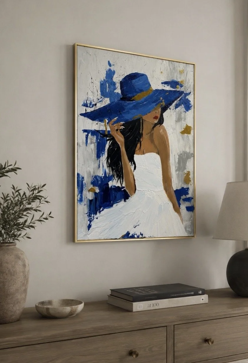 An abstract painting of a woman in a white dress with long black hair, wearing a large blue hat, hanging on a wall above a wooden dresser. The dresser has a potted plant, a small bowl, and a stack of books, with a table lamp on the right.