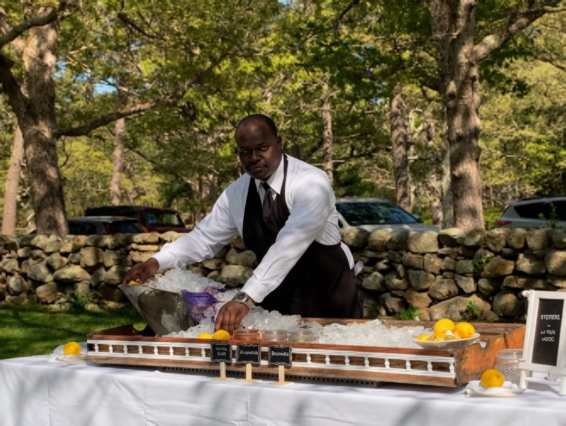 A man dressed in formal attire, including a white shirt and black tie, stands behind a table outdoors, serving oysters, clam from a large container with lemons around it.