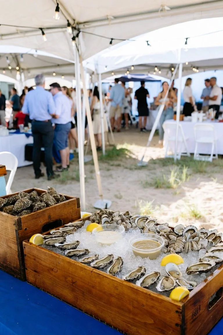 Fresh oysters on ice with lemon wedges and dipping sauce at an outdoor event under white tents with people mingling in the background.